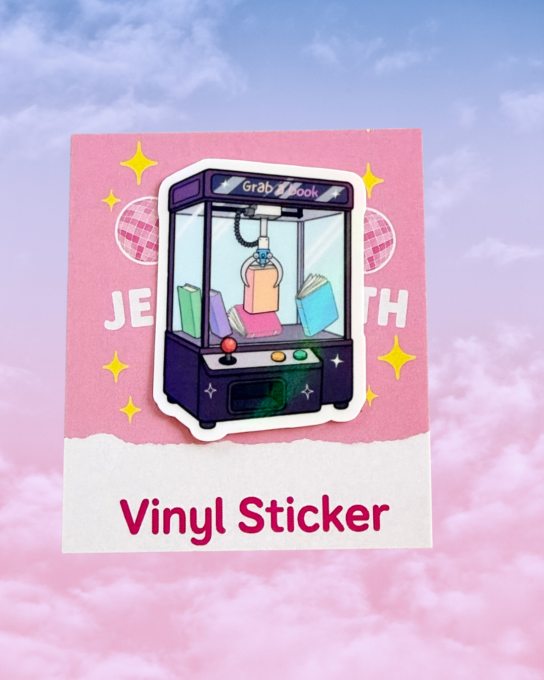 A vinyl sticker featuring an illustrated claw machine with books inside, mounted on a pink card that reads 'Vinyl Sticker' against a pink cloud background.