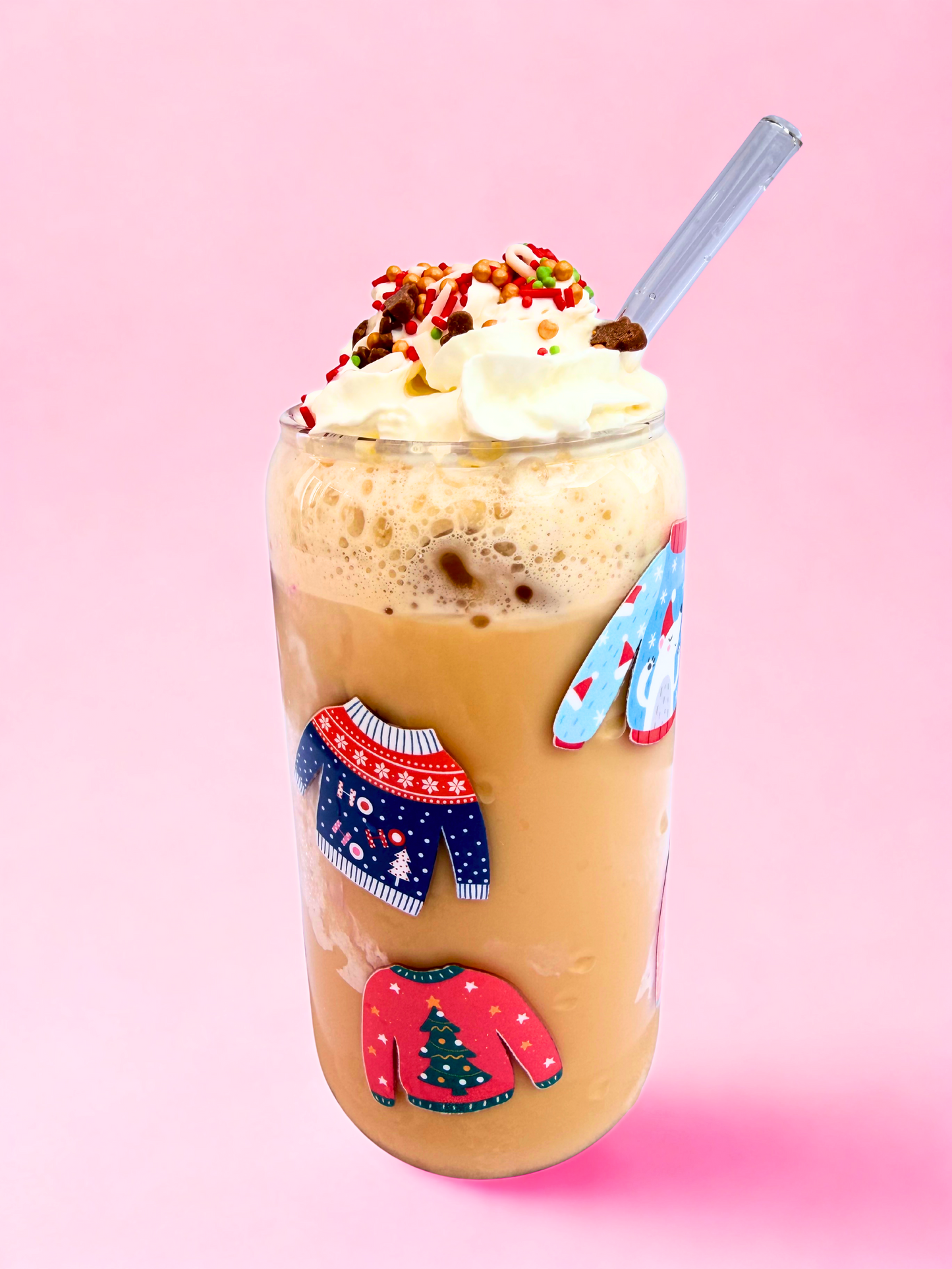 A festive 16oz can glass decorated with colourful Christmas jumper designs, filled with iced coffee topped with whipped cream and sprinkles, with a bamboo lid and glass straw against a pink background.