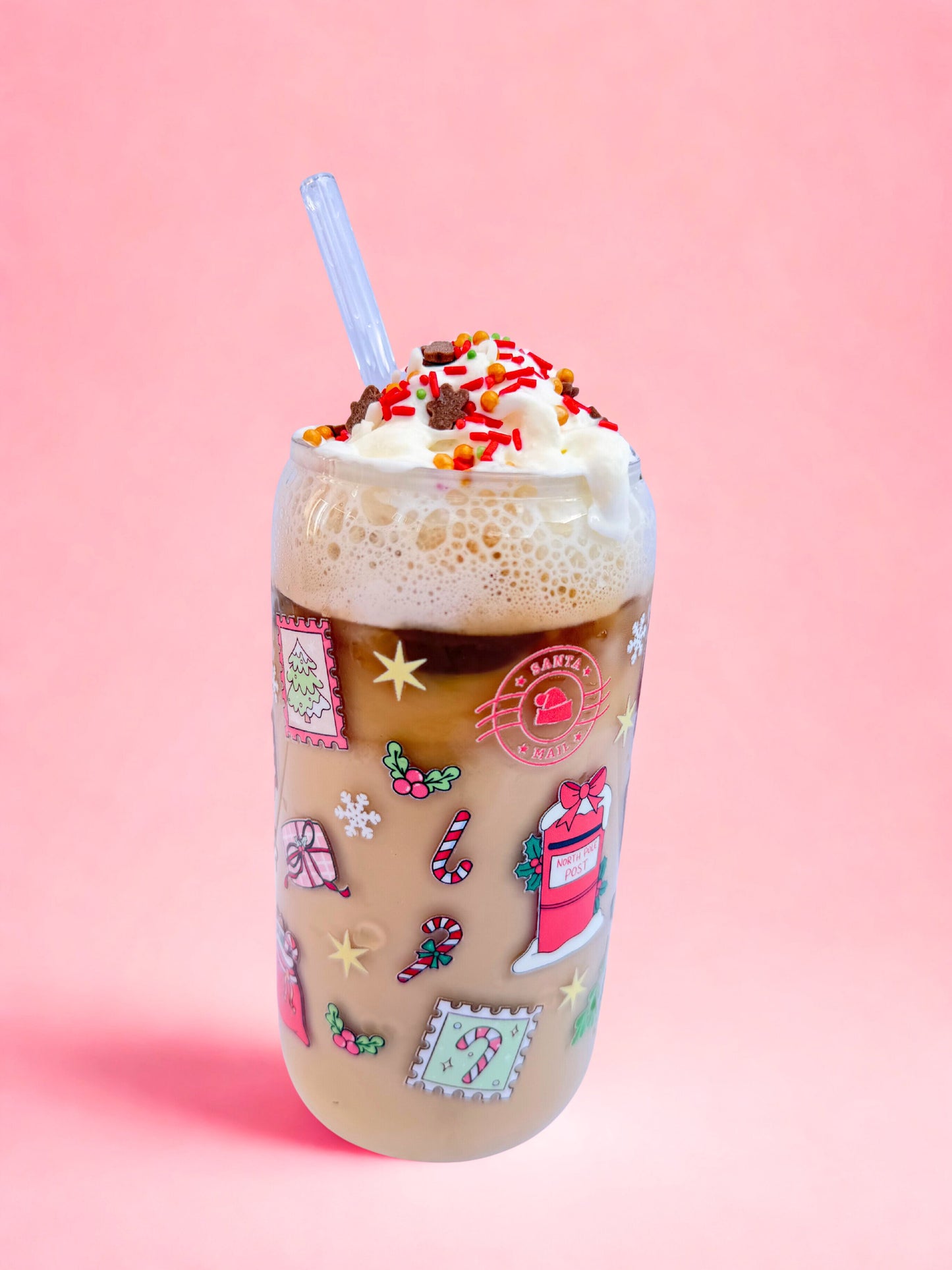 A festive 16oz can glass decorated with Christmas mail designs including Santa’s postbox, candy canes, stamps, and holly, filled with iced coffee topped with whipped cream and sprinkles, against a pink background.