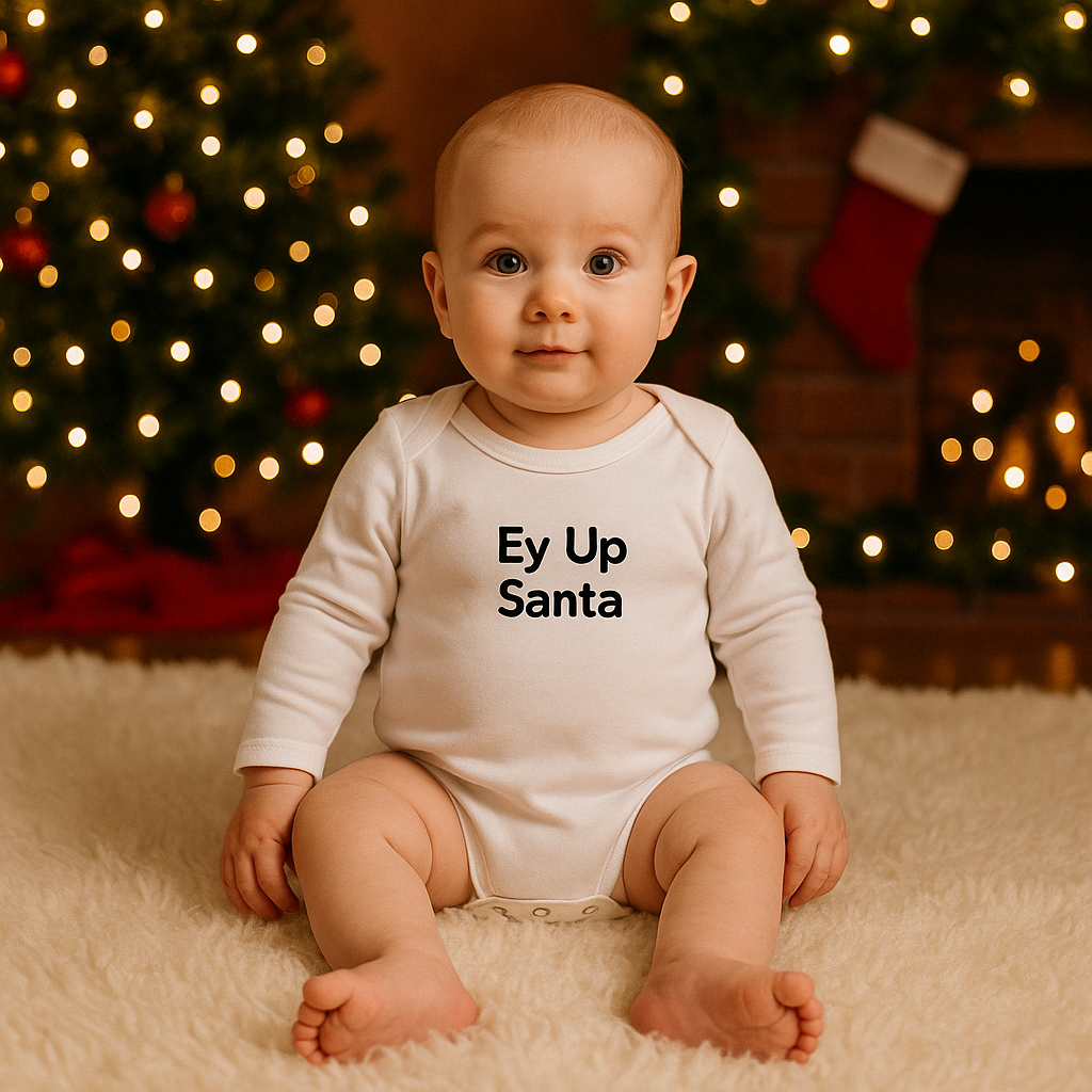 A baby wearing a white long-sleeve bodysuit printed with the words “Ey Up Santa” in bold black letters, sitting on a fluffy rug with Christmas lights and a tree in the background.