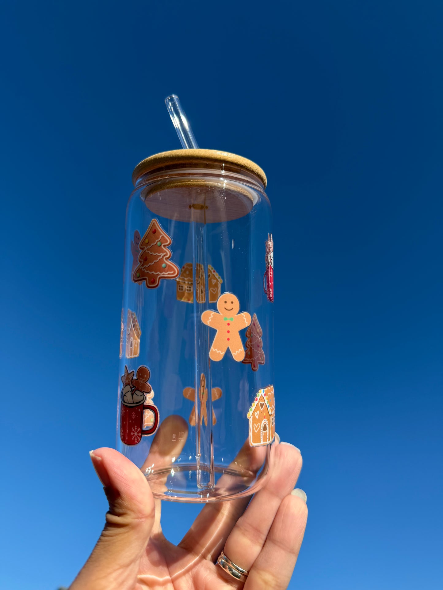 Glass Can Cup with Bamboo lid and glass straw, with images of gingerbread men, gingerbread houses and trees, held up against a blue sky