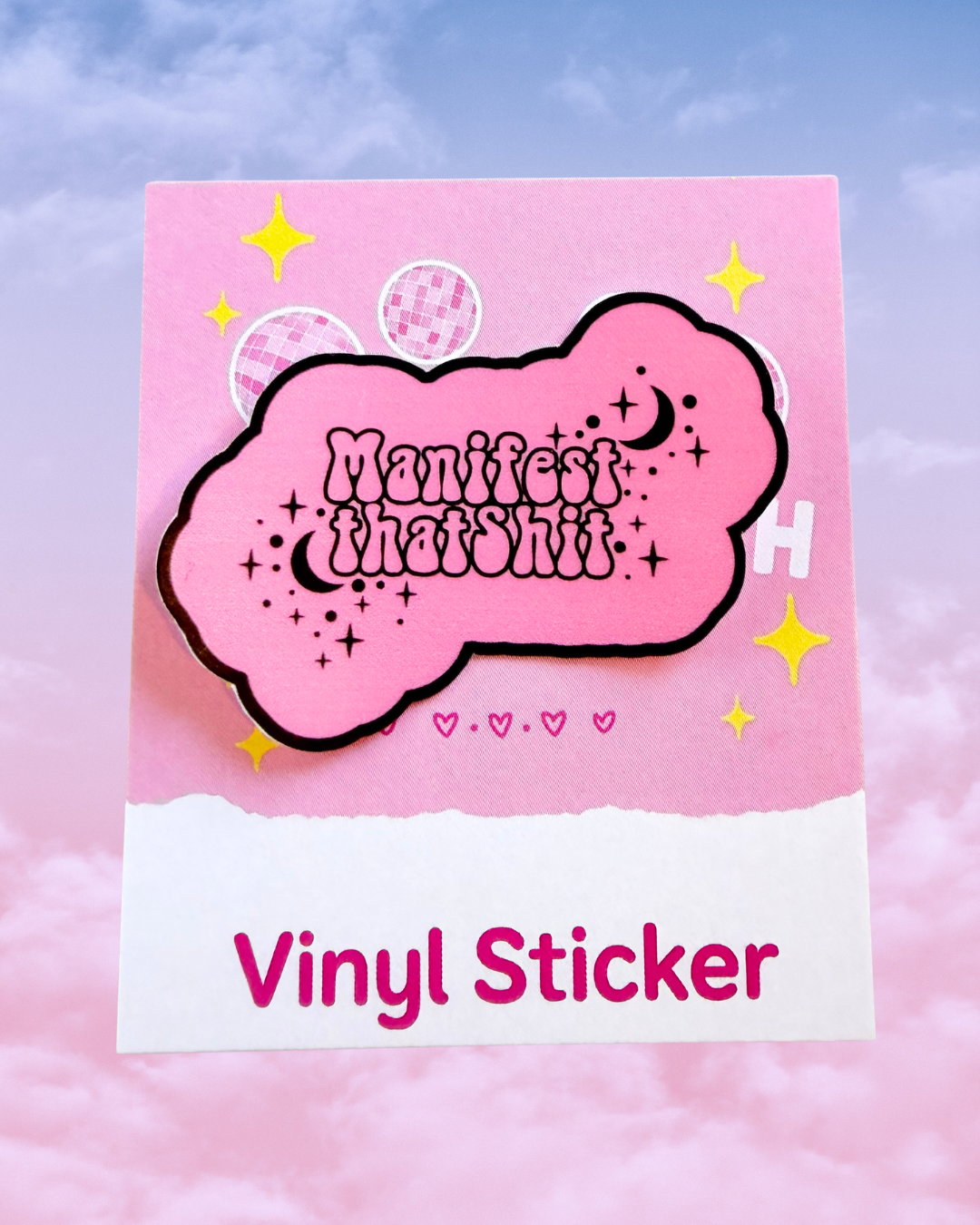 A pink cloud-shaped vinyl sticker with the phrase 'Manifest that sh*t' in stylised lettering, outlined in black, displayed on a pink and white backing card against a soft pink sky background.