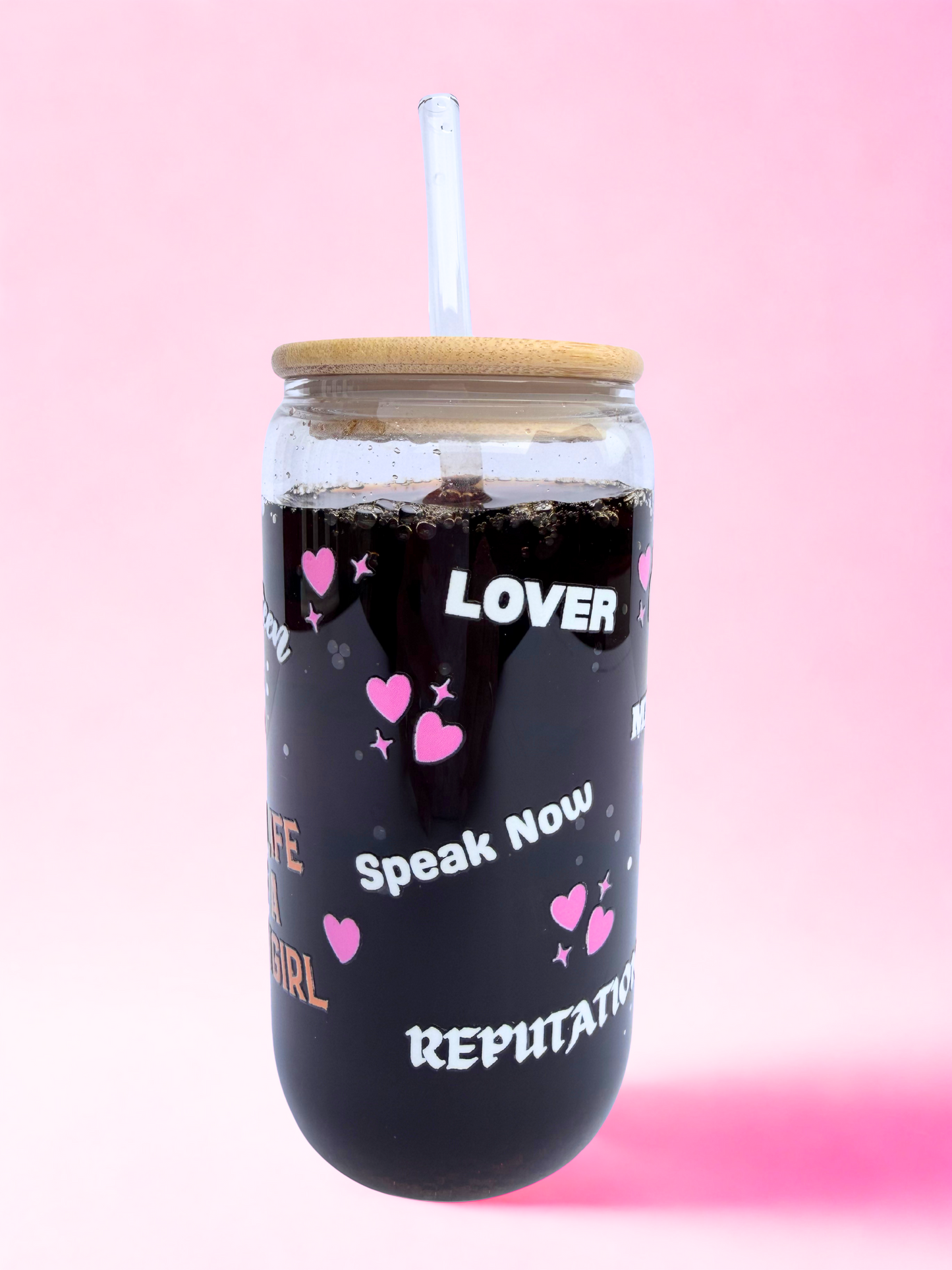 Glass tumbler with bamboo lid and straw, decorated with pink hearts and Taylor Swift Era names, including “The Life of a Showgirl,” on a pink background.