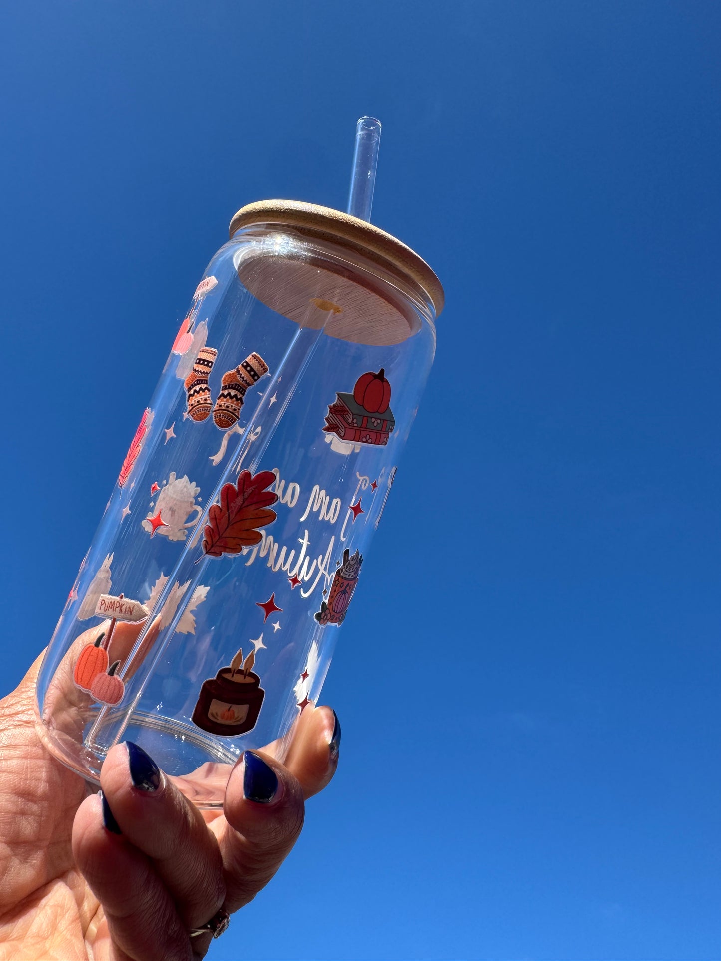 "A clear glass can cup with the words ‘I Am An Autumn’, decorated with cosy seasonal illustrations including socks, a candle, orange bows, a pumpkin-topped book stack, and a pumpkin spice mug – perfect for autumn drinks."