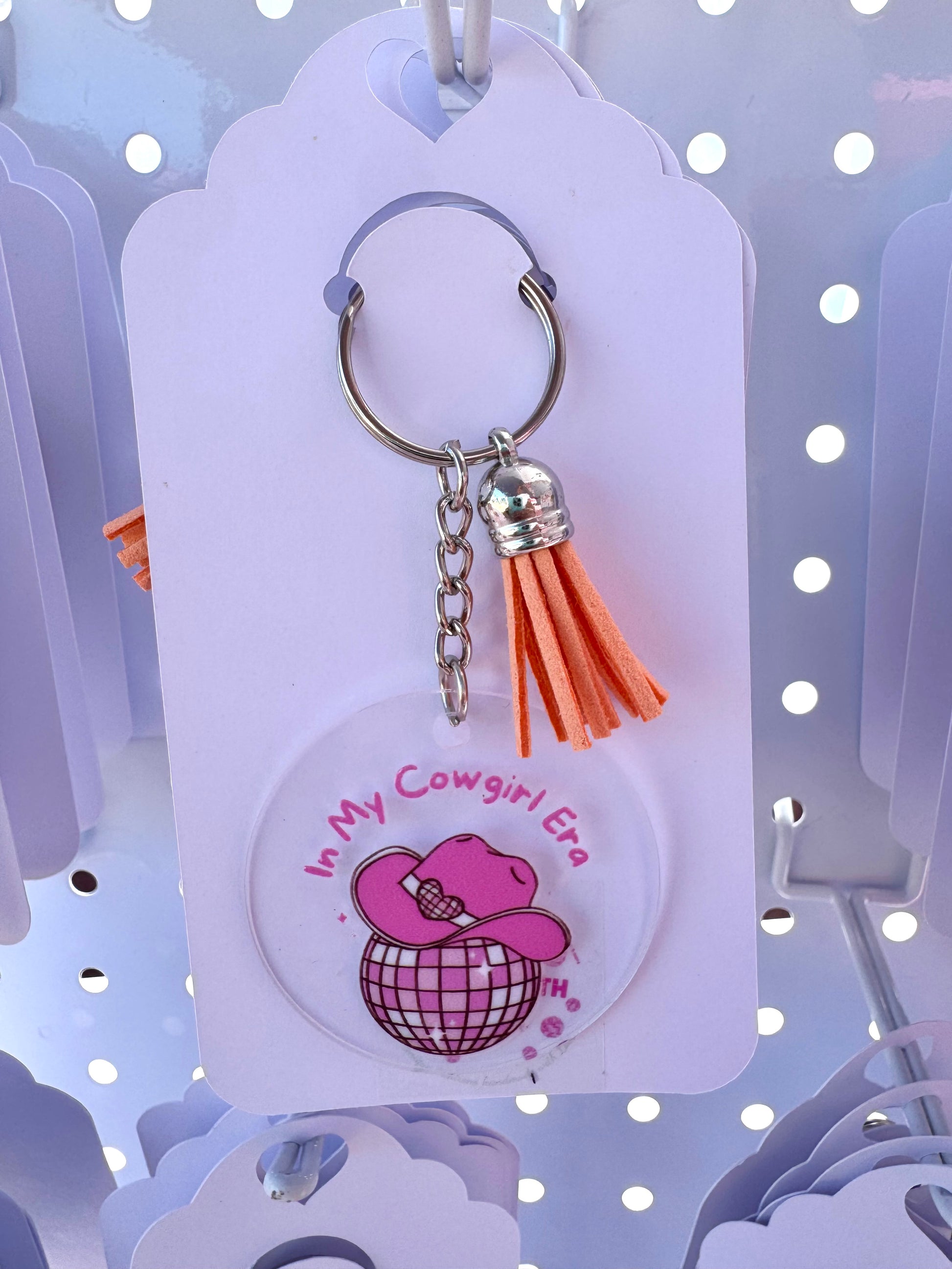 Clear acrylic keychain with a pink cowgirl hat on a disco ball and the words “In My Cowgirl Era,” finished with an orange tassel and silver keyring.