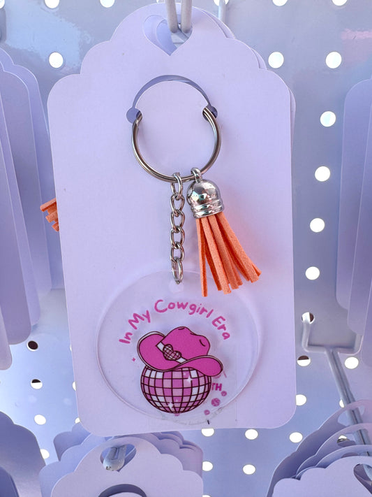 Clear acrylic keychain with a pink cowgirl hat on a disco ball and the words “In My Cowgirl Era,” finished with an orange tassel and silver keyring.