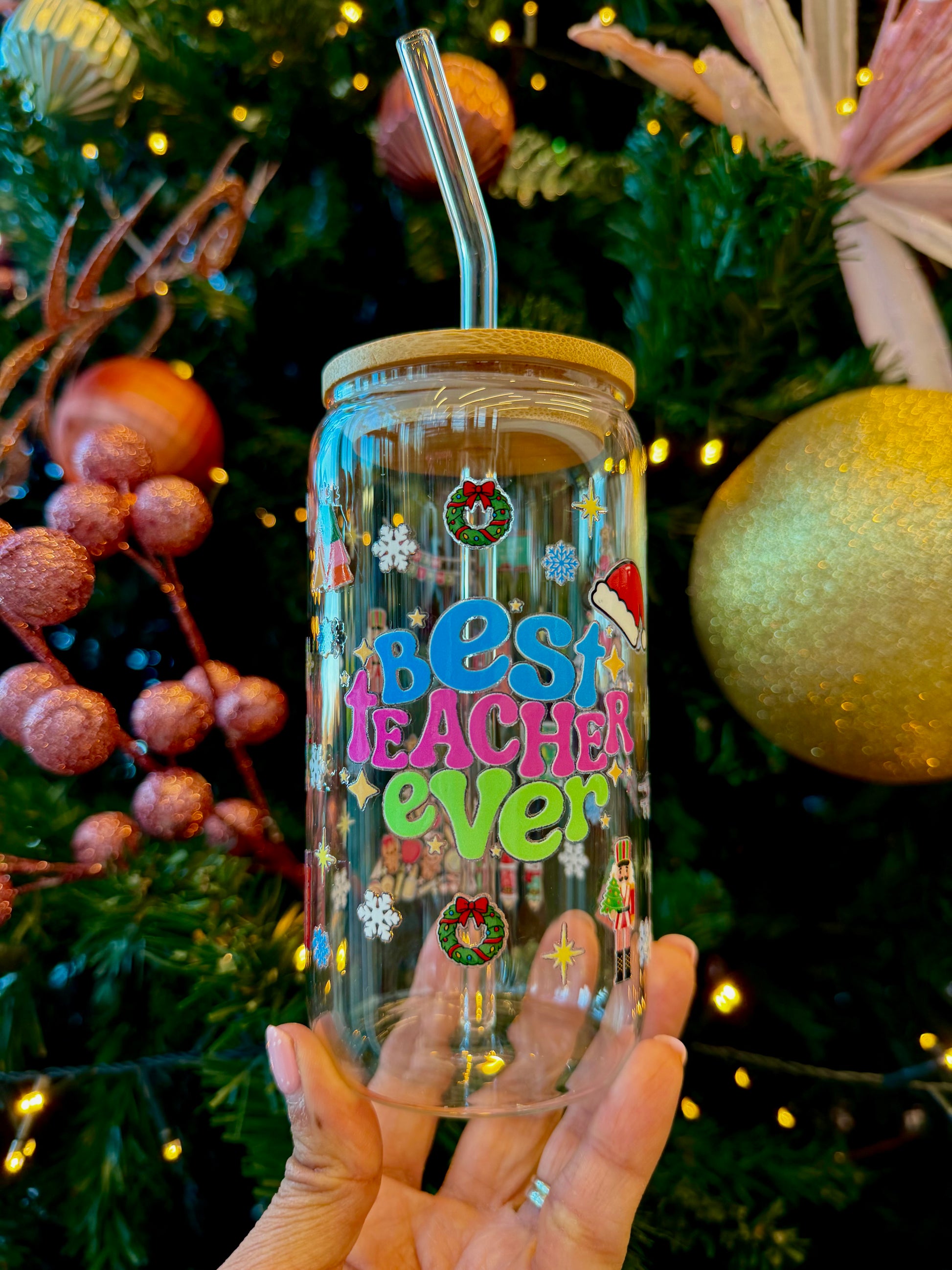 “Christmas-themed glass can cup with colourful ‘Best Teacher Ever’ design, bamboo lid, and glass straw, held in front of a decorated Christmas tree.”