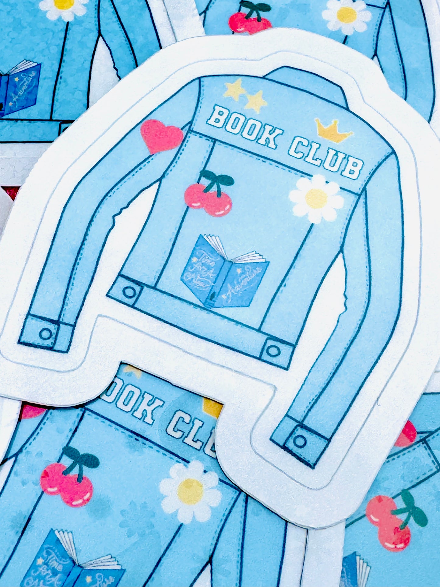 Vinyl sticker featuring a light blue denim jacket with the phrase ‘Book Club’ in white font on the back, along with a flower, book, and heart design, photographed on a flat surface