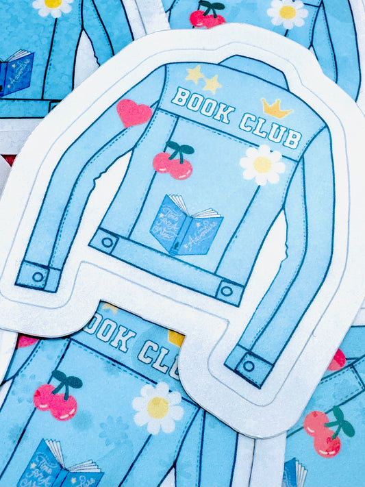 Vinyl sticker featuring a light blue denim jacket with the phrase ‘Book Club’ in white font on the back, along with a flower, book, and heart design, photographed on a flat surface