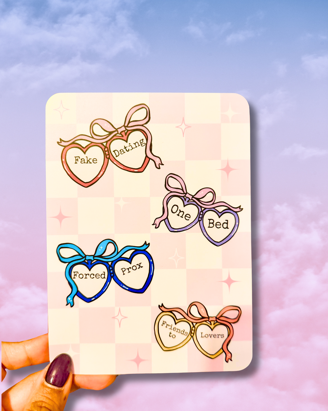 A hand with dark purple nail polish holds a kindle insert featuring five illustrated pairs of hearts with bows and words like 'Fake Dating,' 'One Bed,' 'Forced Proximity,' 'Friends to Lovers,' and 'Fake Love.' The card is shown against a pastel sky background with clouds.
