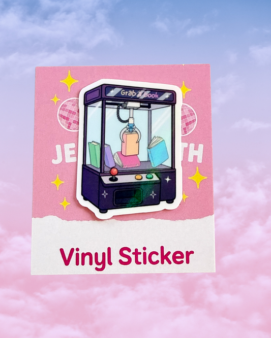 A vinyl sticker featuring an illustrated claw machine with books inside, mounted on a pink card that reads 'Vinyl Sticker' against a pink cloud background.
