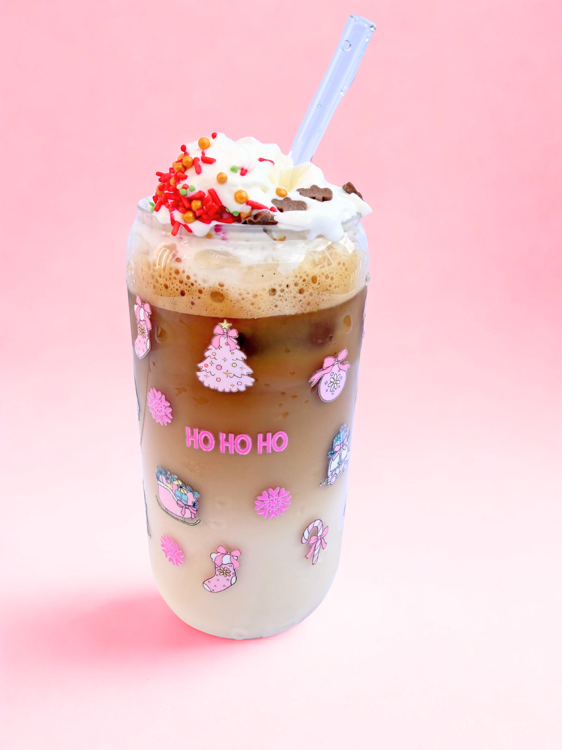 Glass can cup with pink festive image on it, such as ho ho ho, christmas tree and baubles. It is on a pink background
