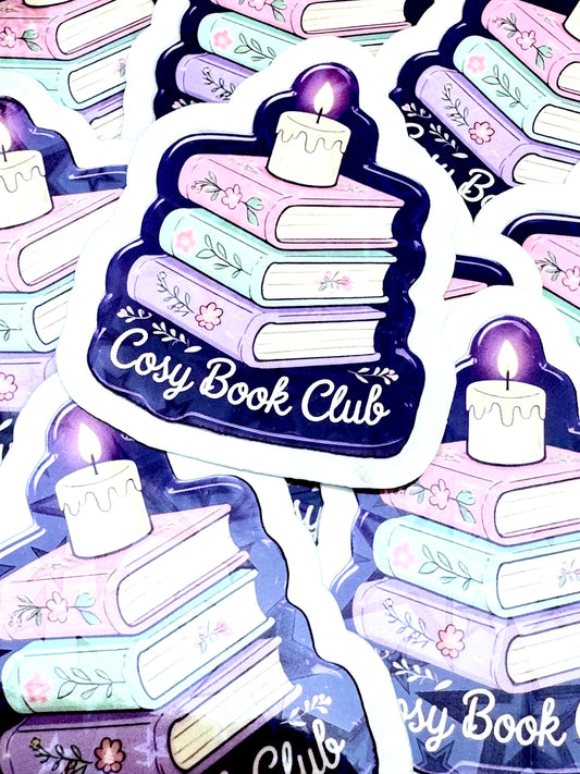 Purple vinyl sticker with a stack of books topped by a candle, and the phrase ‘Cosy Book Club’ in white font, photographed on a flat surface.
