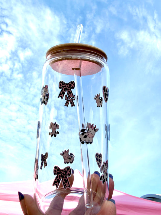 Clear glass can cup with cute pink cow print bows and cartoon cows, featuring a bamboo lid and clear glass straw, held against a blue sky.