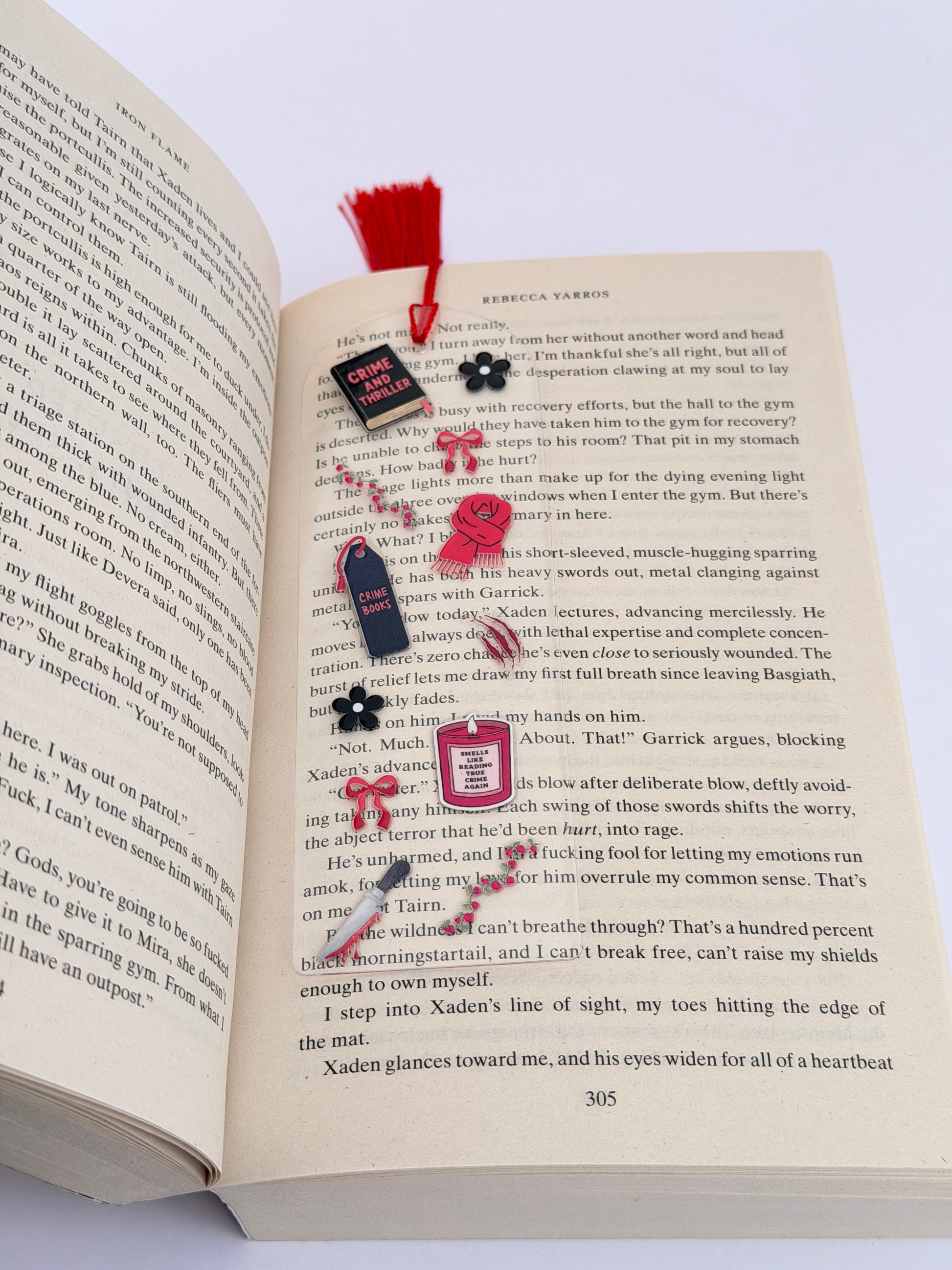 A clear acrylic bookmark featuring red and black crime-themed illustrations, including mini books, a candle, and a knife design, lying open on a thriller novel with a red tassel at the top.