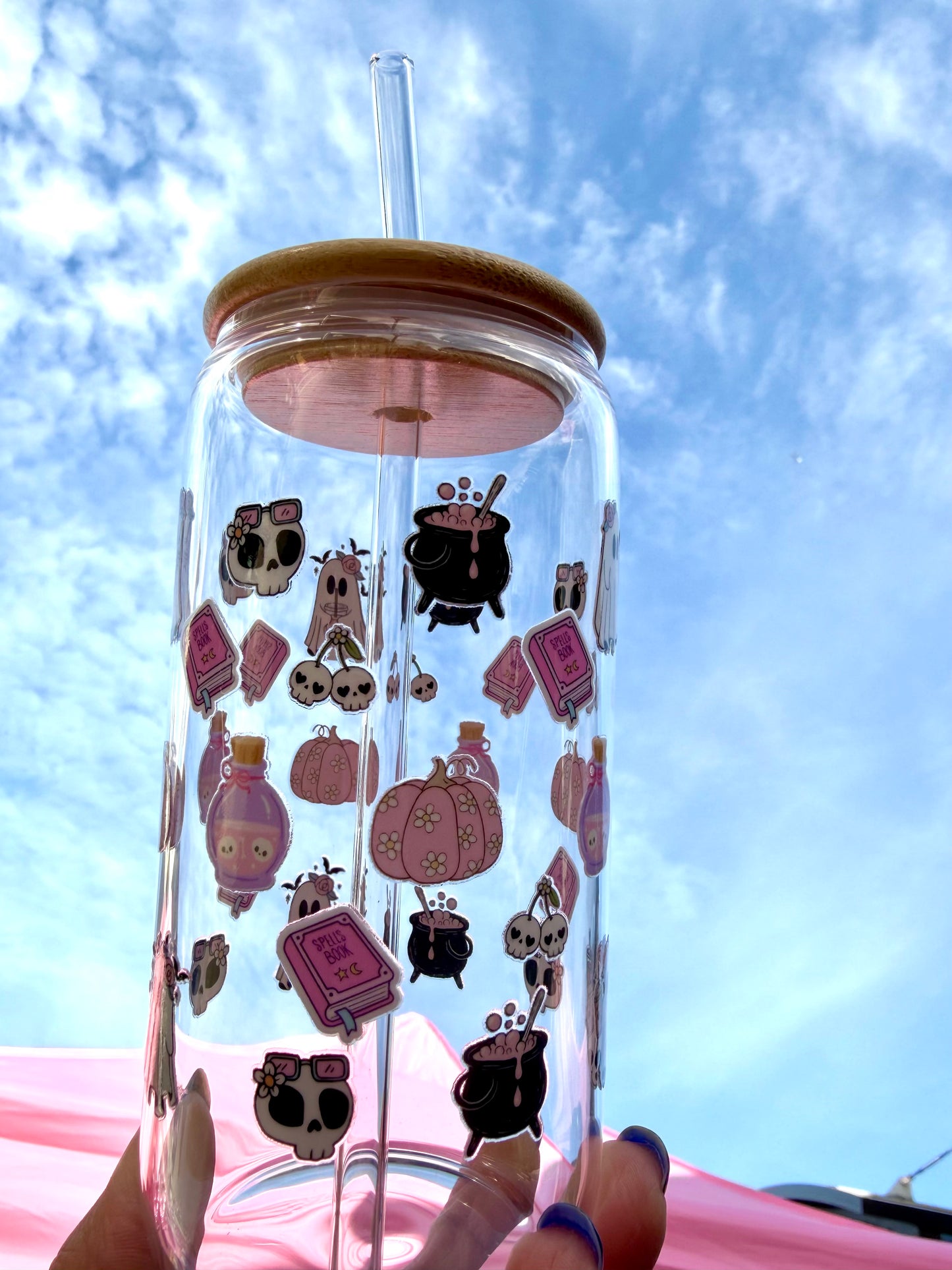 Clear glass can cup with pink witchy designs — skulls, cauldrons, and potions — plus bamboo lid and glass straw.