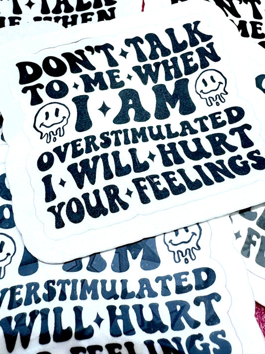 White vinyl sticker with the phrase ‘Don’t talk to me when I’m overstimulated, I will hurt your feelings’ in black font, photographed on a flat surface.