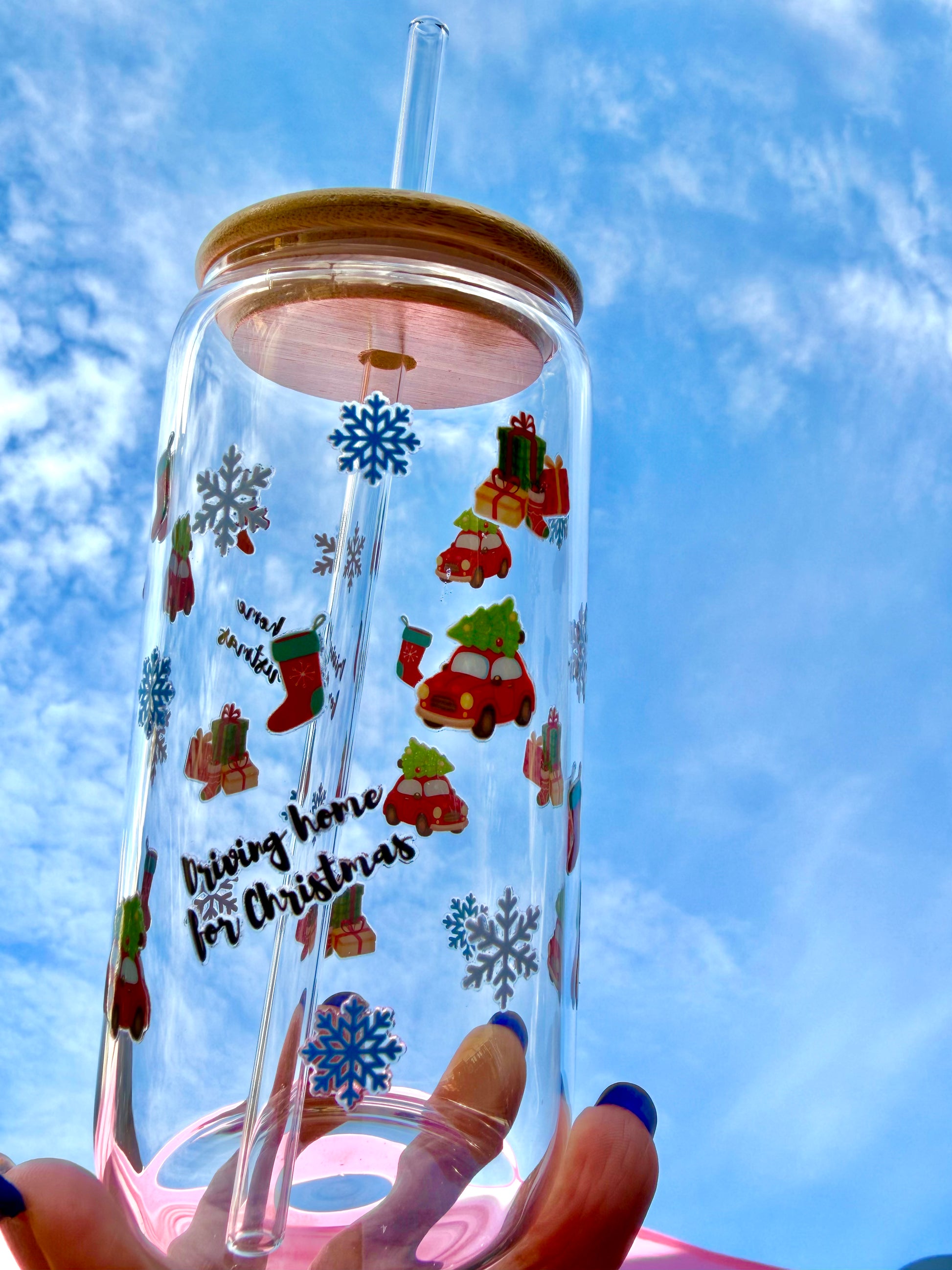 A reusable “Driving Home for Christmas” glass cup with bamboo lid and glass straw, decorated with red cars carrying presents, stockings, and snowflakes, held against a bright blue sky.