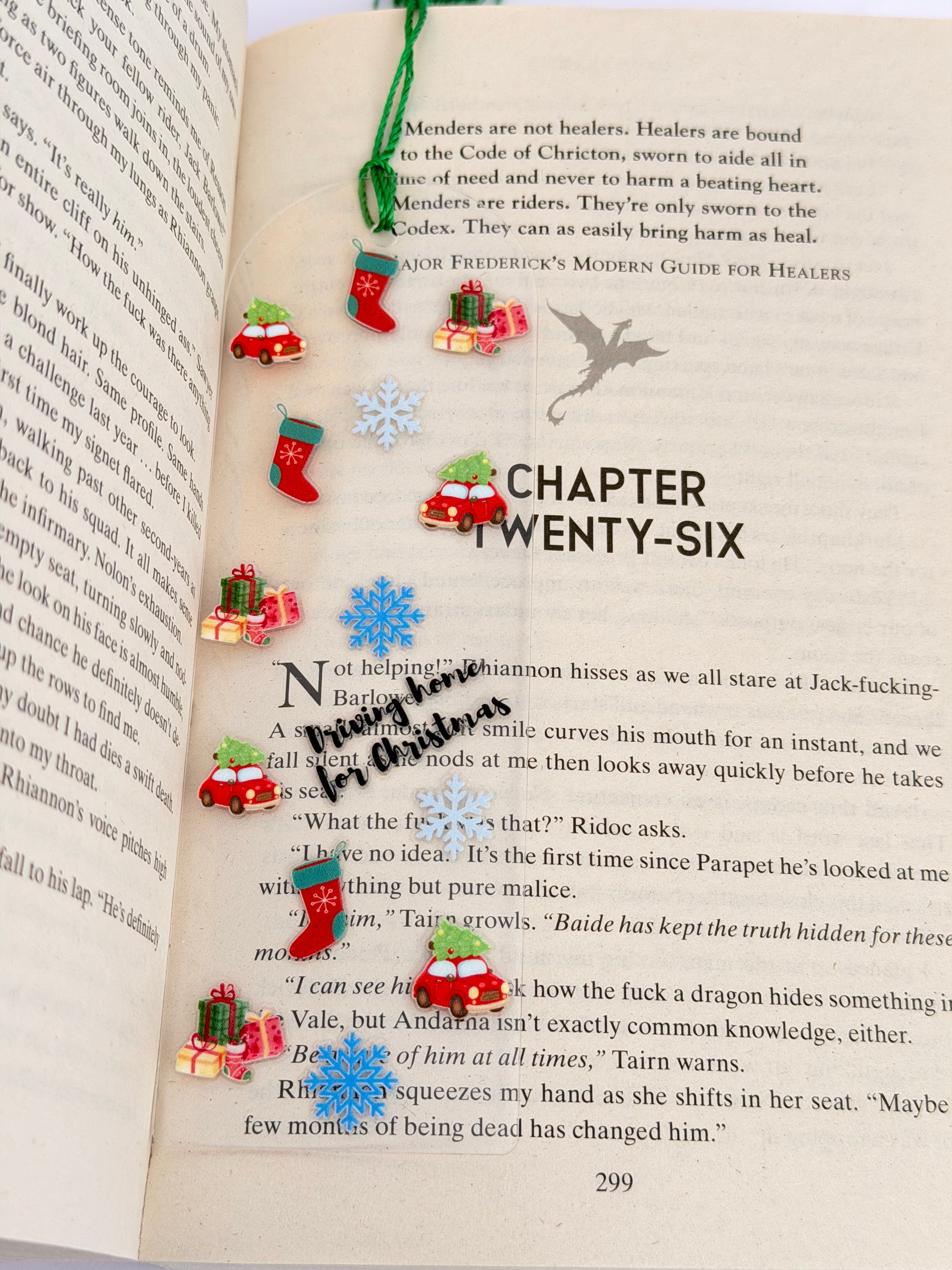 A festive acrylic bookmark decorated with Christmas charms including red cars carrying trees, stockings, snowflakes, and presents, laid open across a book with the text “Driving Home for Christmas.”