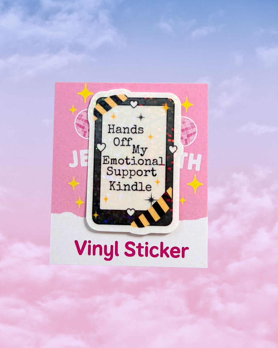 A vinyl sticker product shown on a pink backing card against a pastel sky background. The sticker features a e-reader design with the text 'Hands Off My Emotional Support Kindle' in black on a cream center, surrounded by glittery black and orange diagonal hazard stripes, small white hearts, and sparkles.