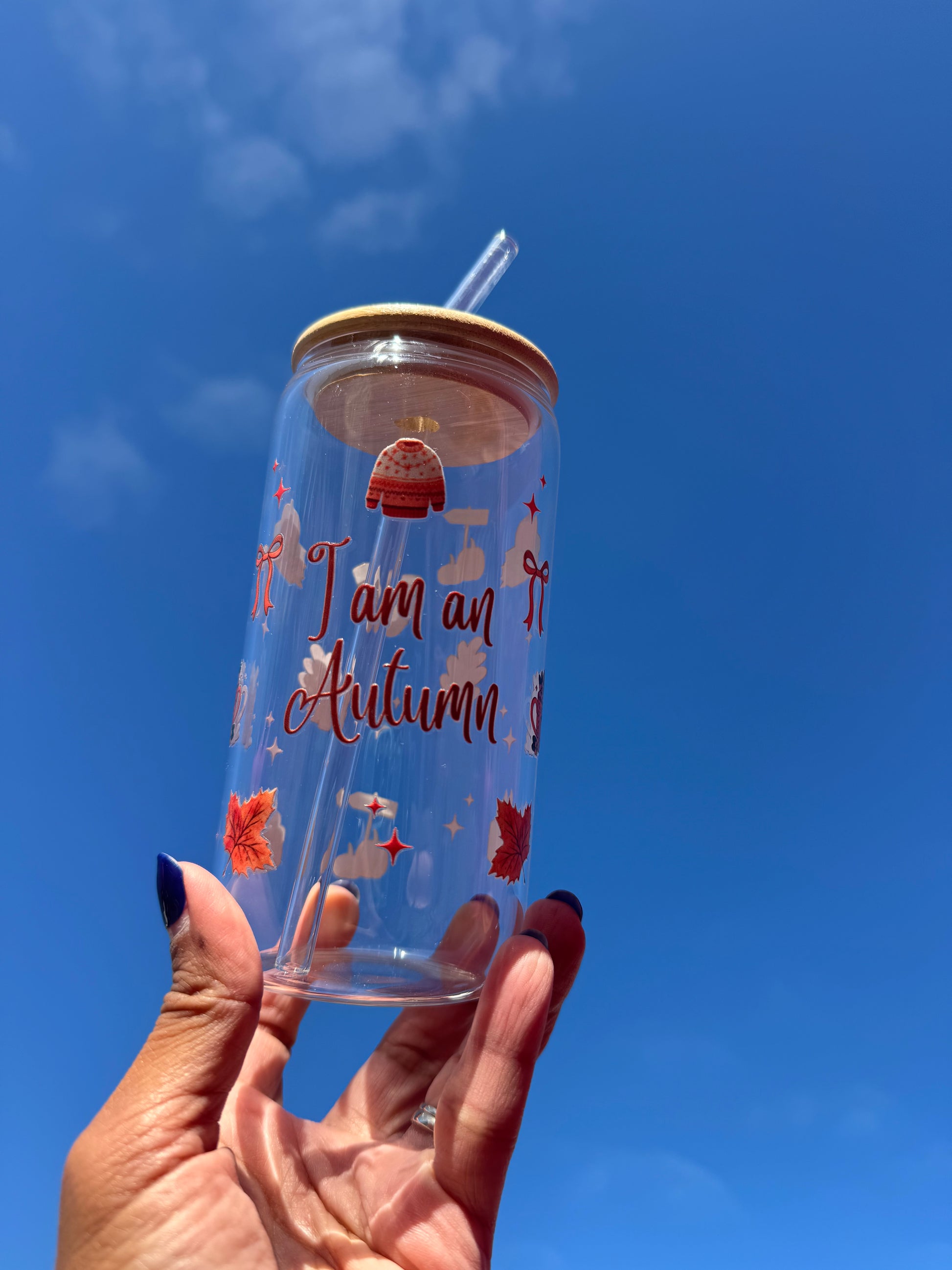"A clear glass can cup with the words ‘I Am An Autumn’, decorated with cosy seasonal illustrations including socks, a candle, orange bows, a pumpkin-topped book stack, and a pumpkin spice mug – perfect for autumn drinks."