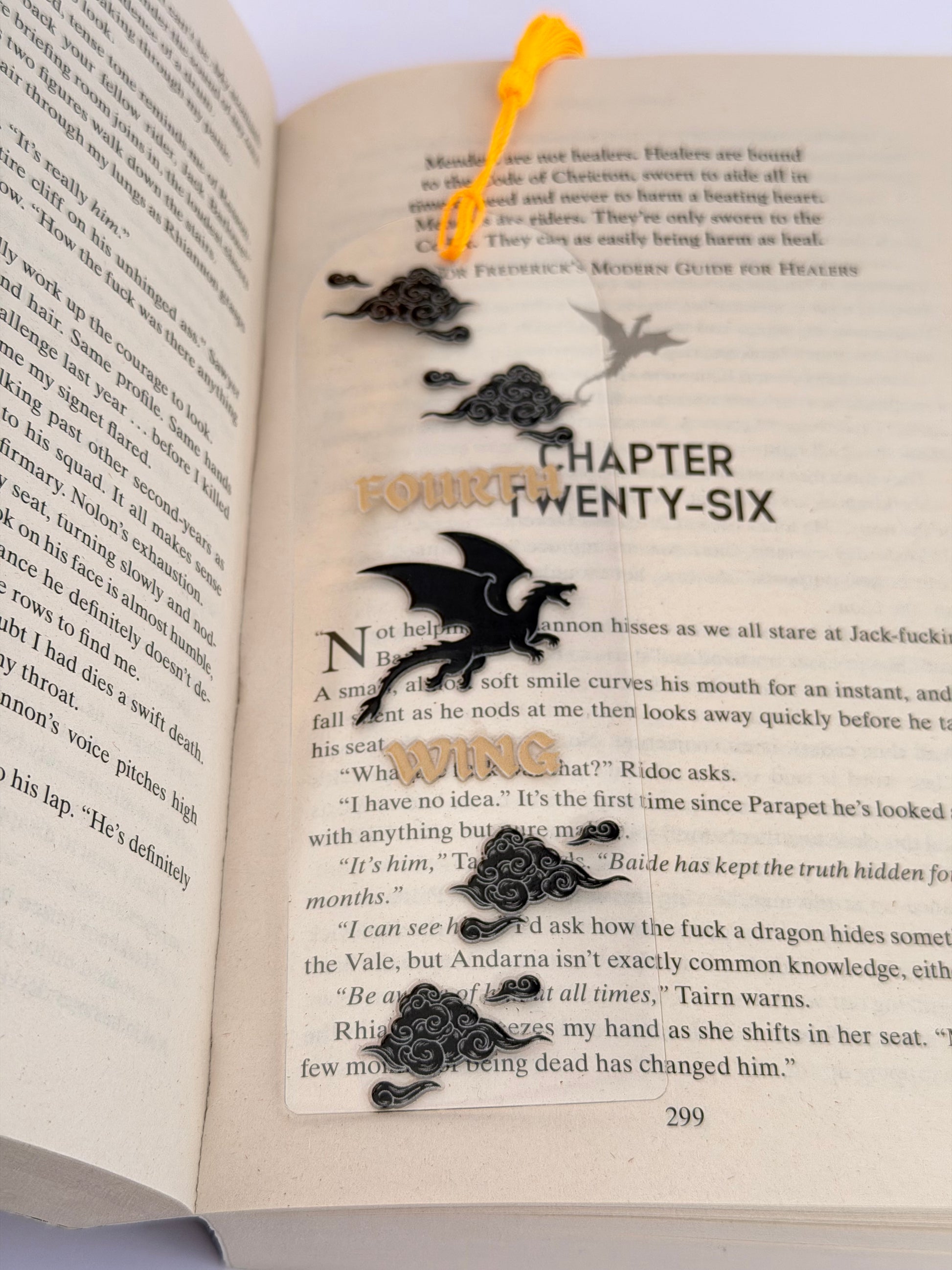 A clear acrylic bookmark featuring black clouds, a dragon silhouette, and gold text reading “Fourth Wing,” resting on an open fantasy book with a yellow tassel at the top.
