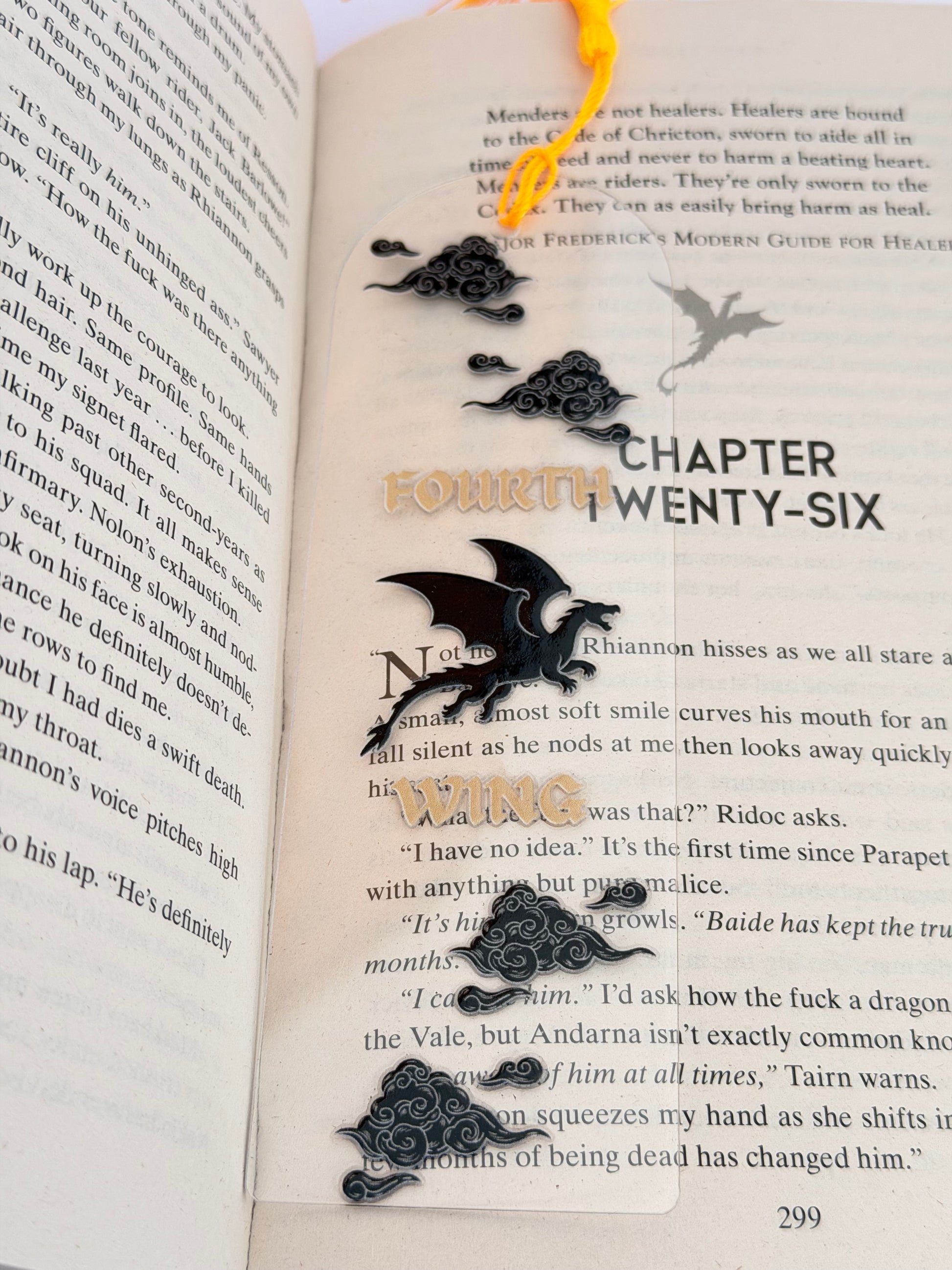 Close-up of The Fourth Wing Acrylic Bookmark showing dragon and cloud illustrations with metallic gold lettering, placed on an open page.