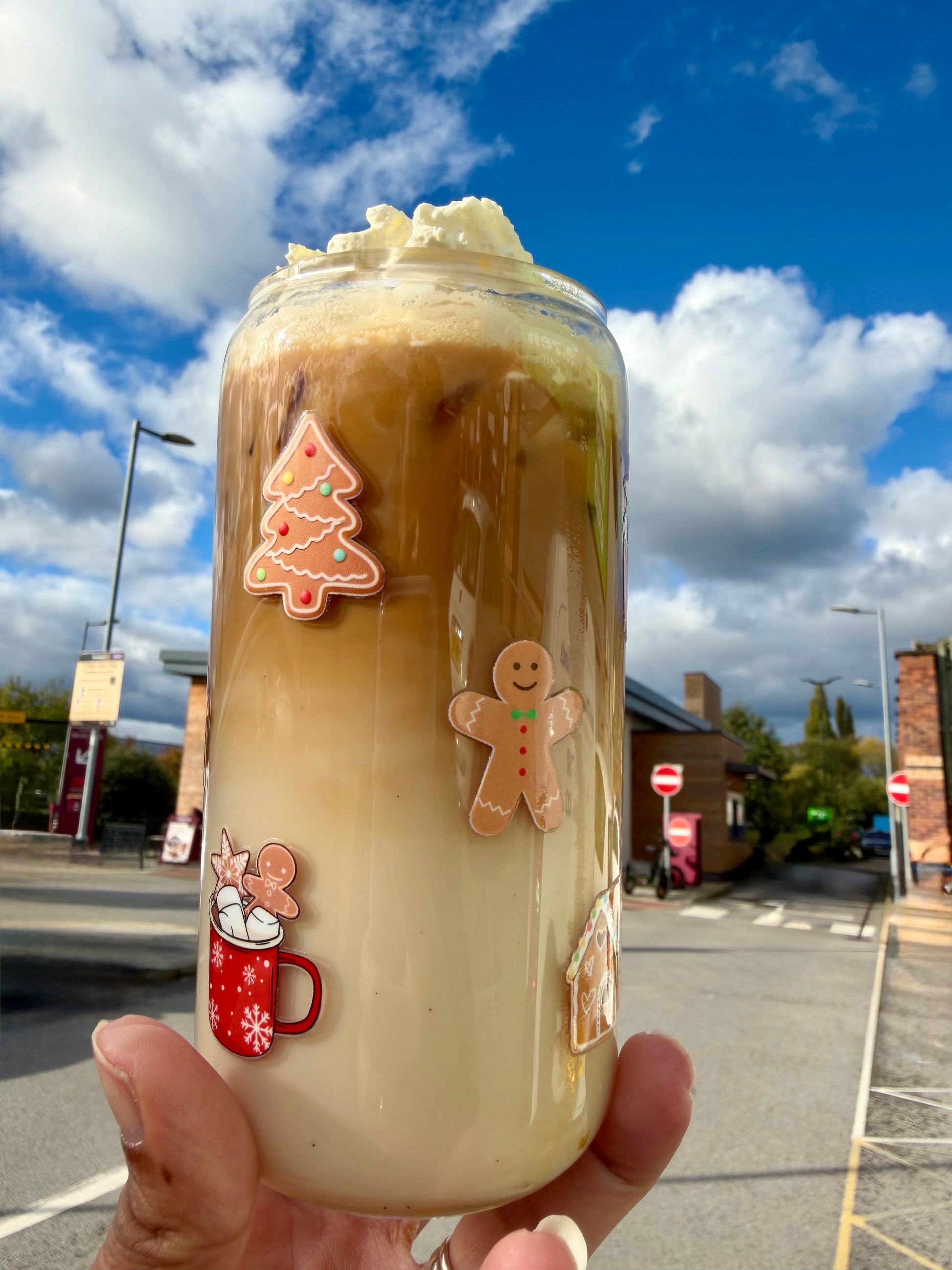 A reusable glass cup with a bamboo lid featuring gingerbread, Christmas tree, and festive mug designs, filled with an iced latte topped with whipped cream, photographed under a bright blue sky.