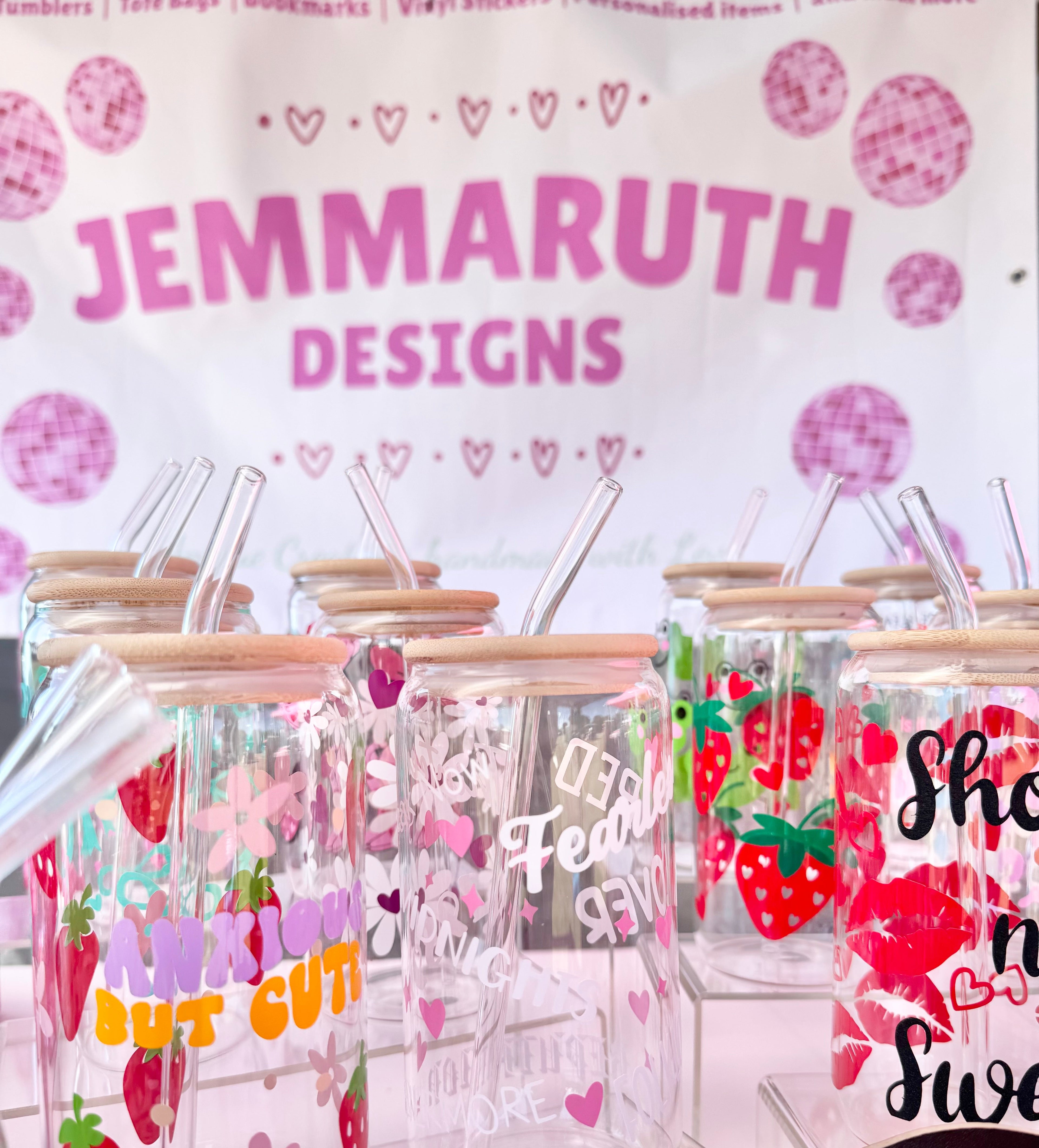 Glass can cups with lots of designs on them such as strawberry designs and text on a stand, branded 'Jemmaruth Designs'.