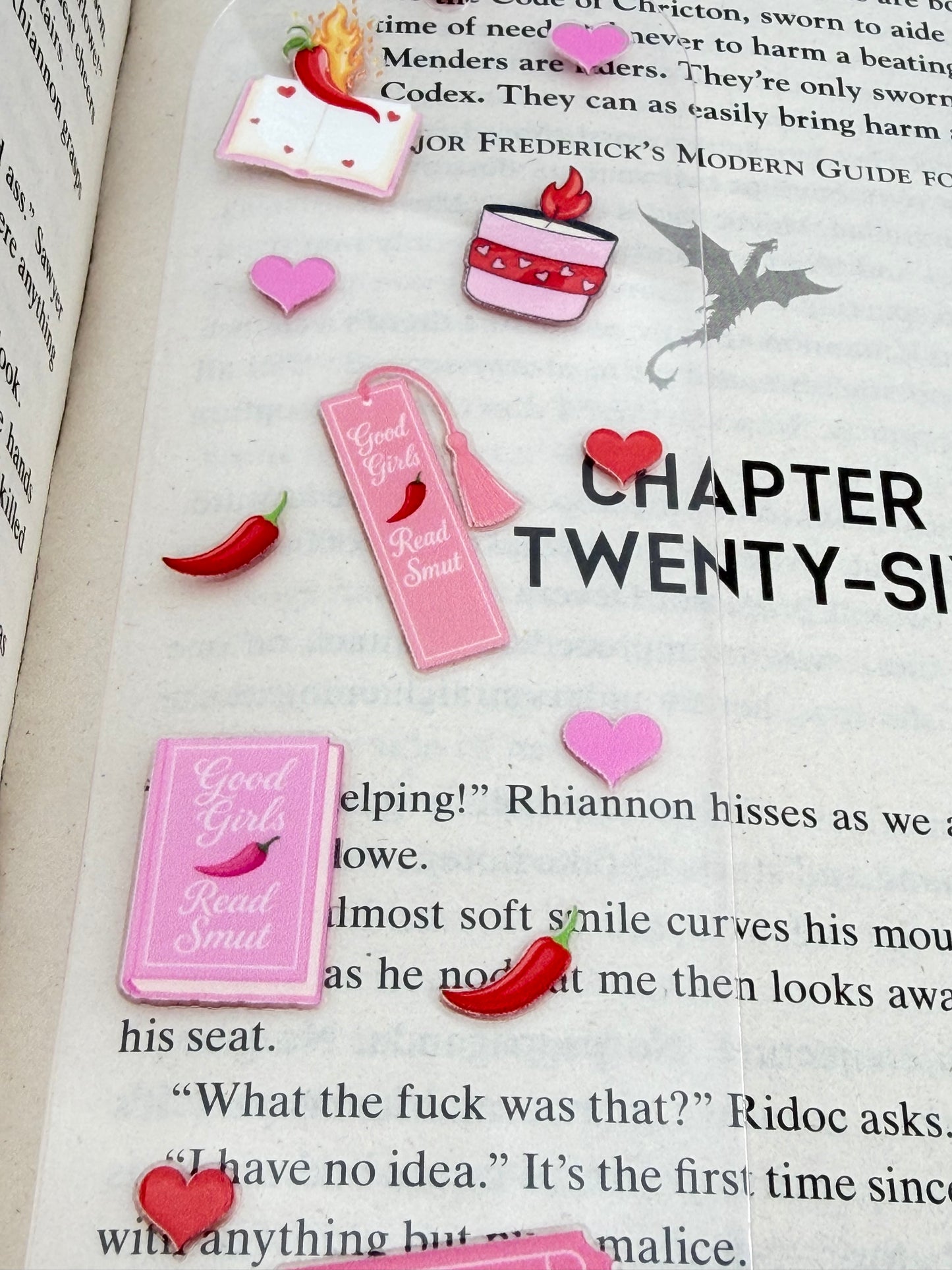 A close up of a clear acrylic bookmark with pink and red spicy book-themed designs, including hearts, books, and chilli peppers, lying across an open book with a pink tassel at the top.