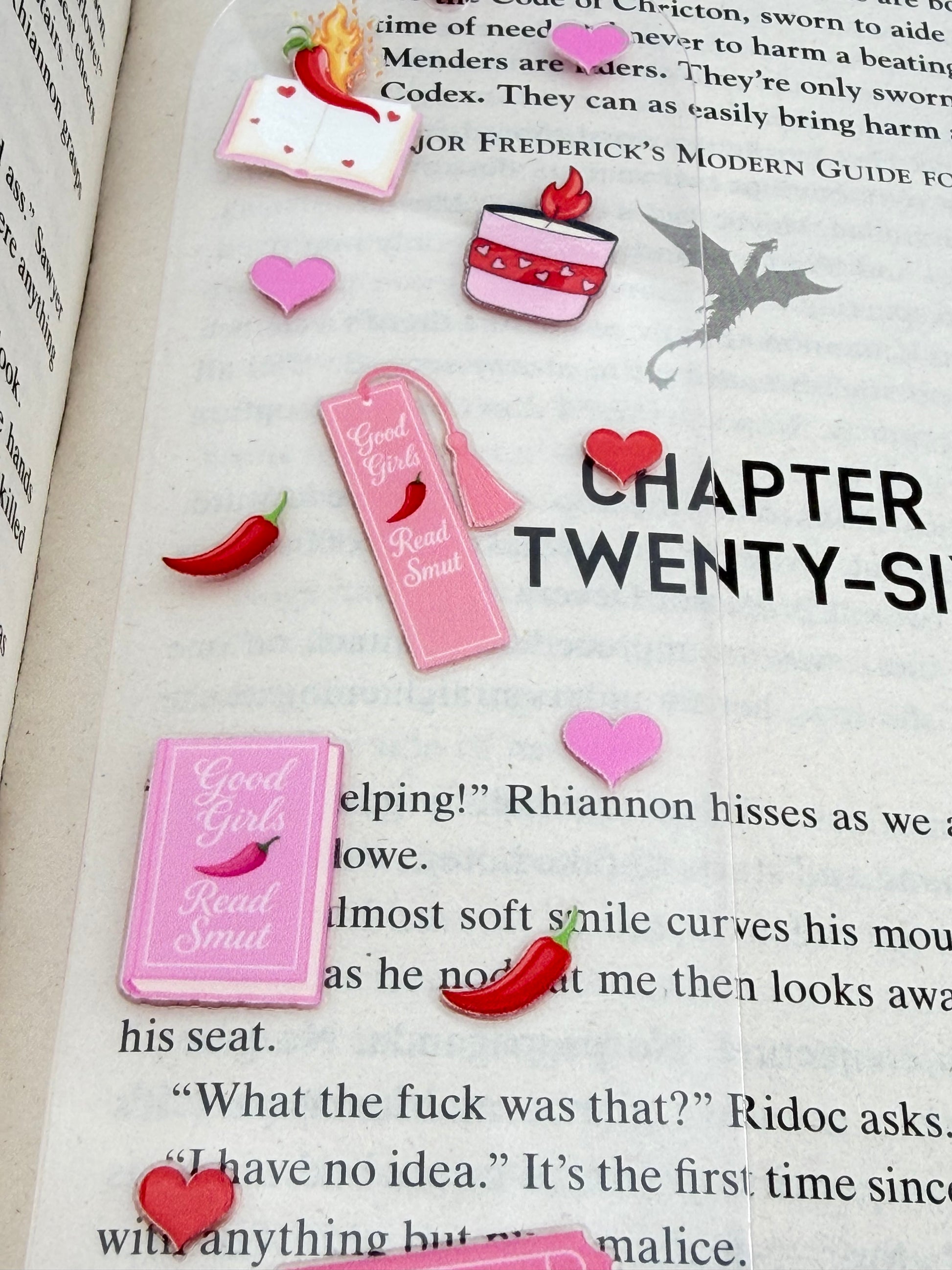 A close up of a clear acrylic bookmark with pink and red spicy book-themed designs, including hearts, books, and chilli peppers, lying across an open book with a pink tassel at the top.