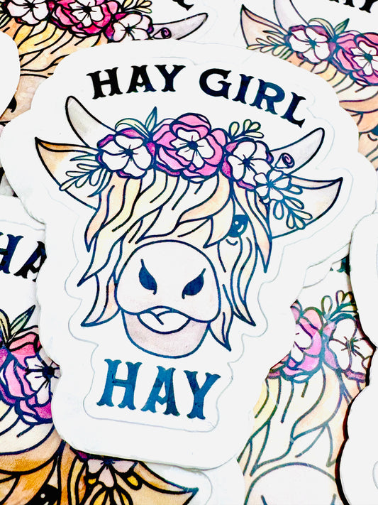 Vinyl sticker featuring a Highland cow with flowers in its hair and the phrase ‘HAY Girl HAY’ in black font, photographed on a flat surface.
