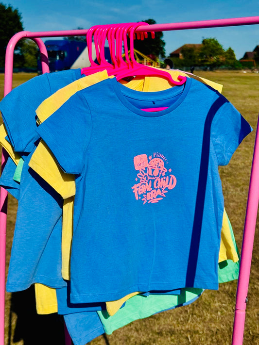 Blue children’s t-shirt with ‘In My Feral Child Era’ in bold orange font, hanging on a pink hanger on a pink rail, photographed outdoors in the countryside.