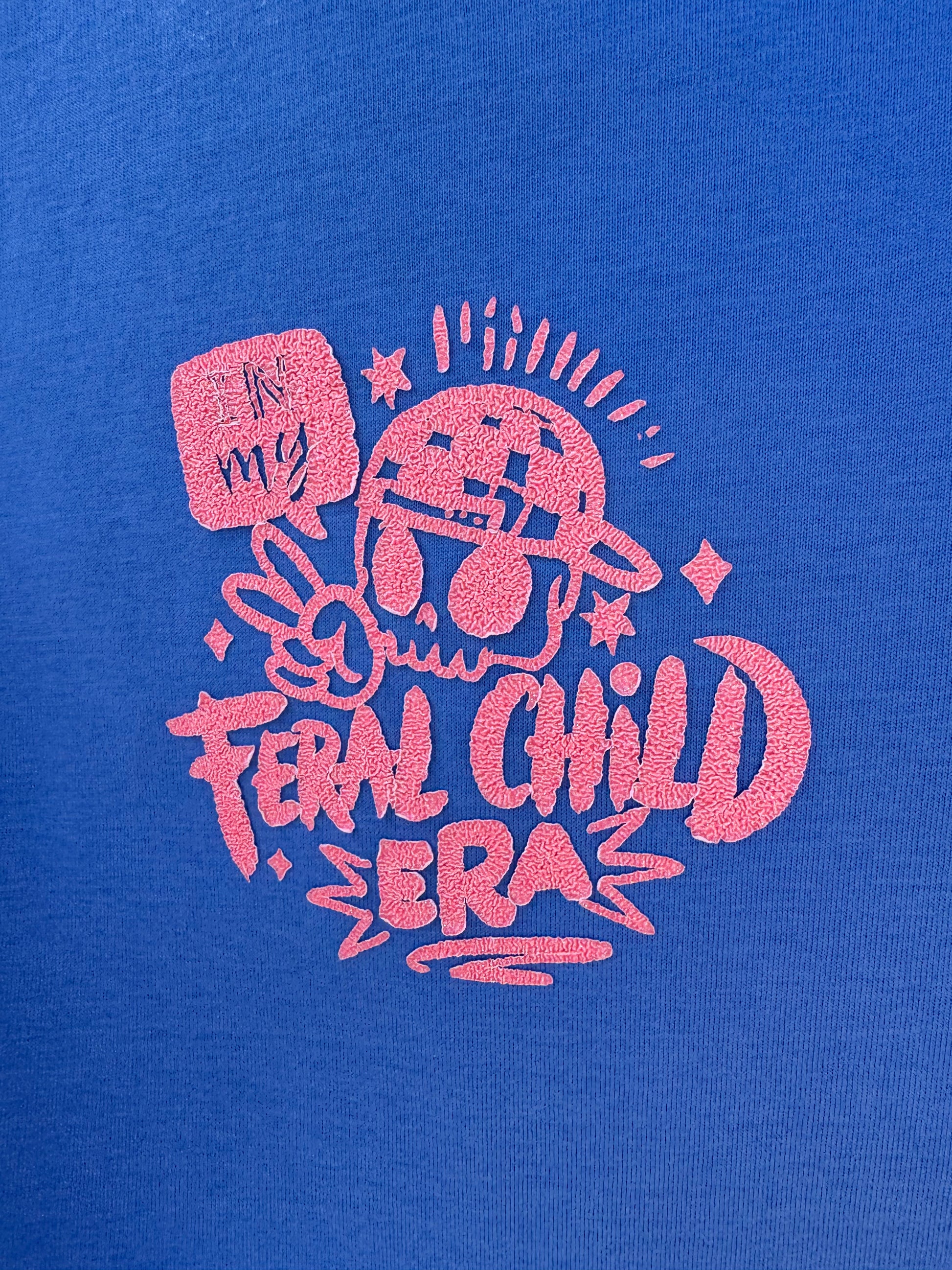 Close up of a blue T-shirt with orange logo saying in my feral child era with a peace sign and a face with a backward cap