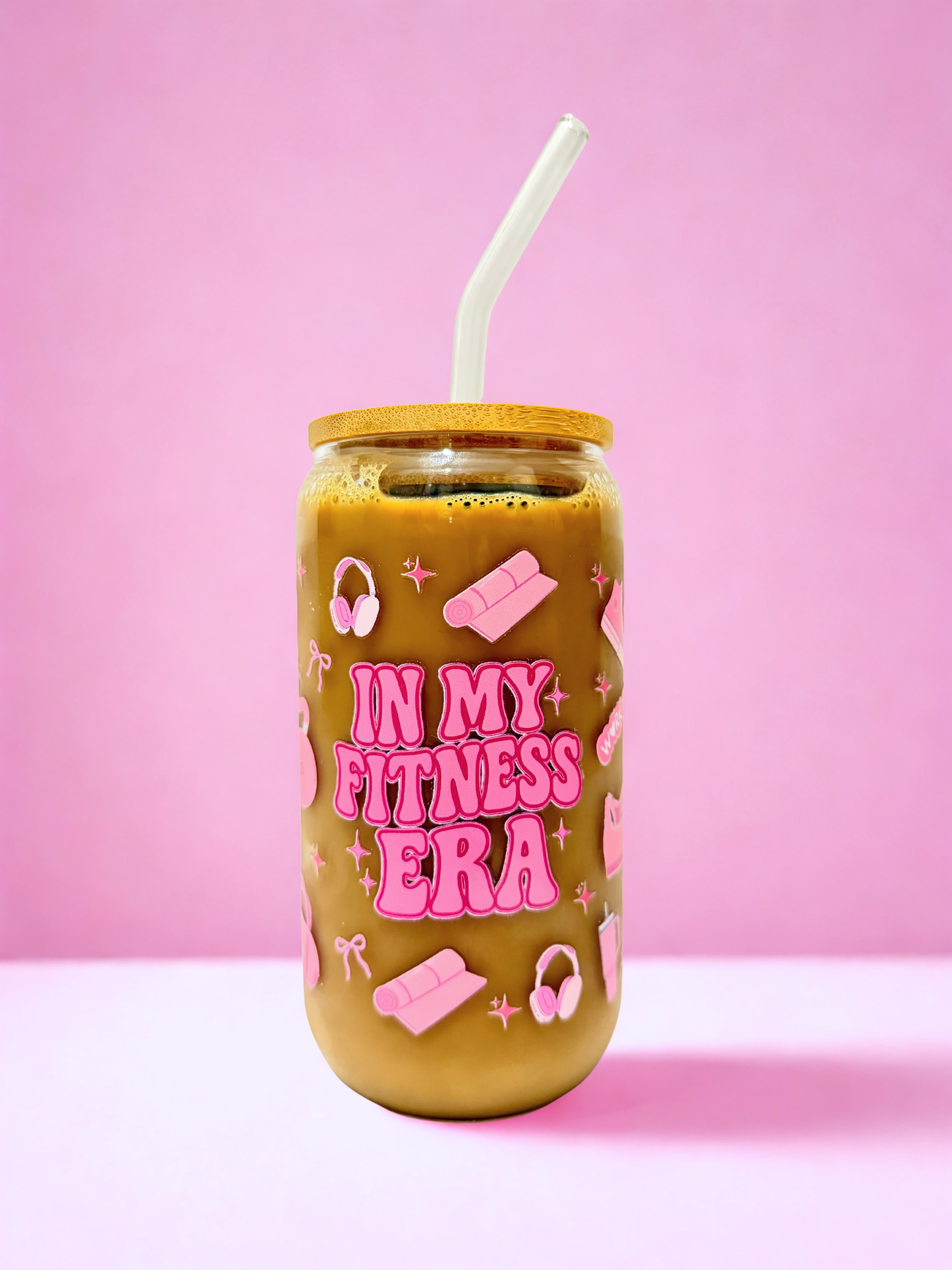 “A 16oz glass can cup with bamboo lid and clear glass straw, decorated with pink fitness icons and the phrase ‘In My Fitness Era,’ photographed against a pink background.”