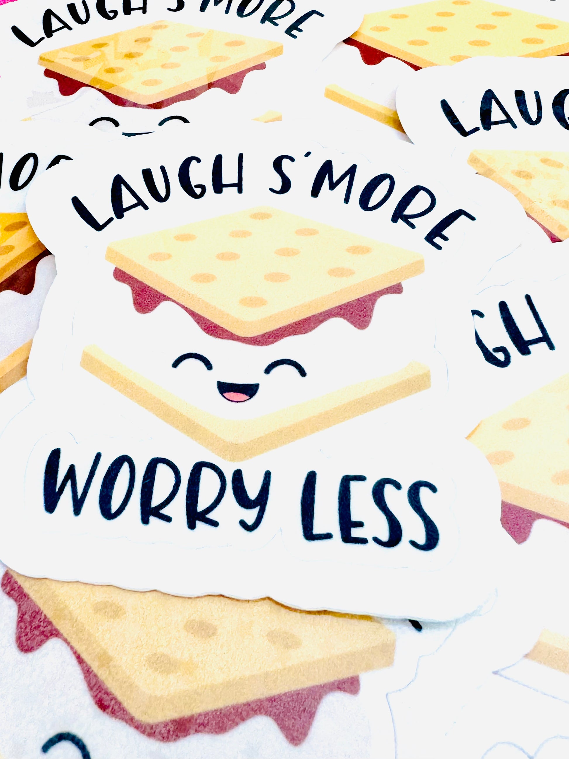 Vinyl sticker with black lettering that says ‘Laugh S’more’ at the top and ‘Worry Less’ at the bottom, featuring a smiley-faced s’more illustration in the center, photographed on a flat surface.
