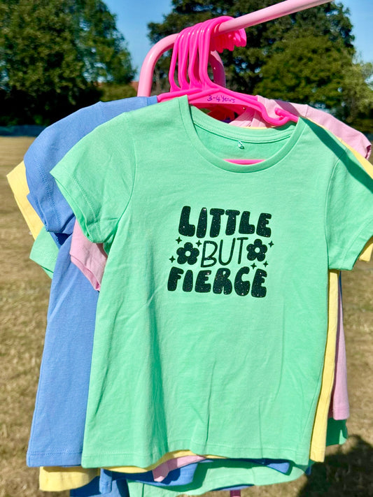 Green t-shirt with 'LITTLE BUT FIERCE' text on a hanger outdoors.
