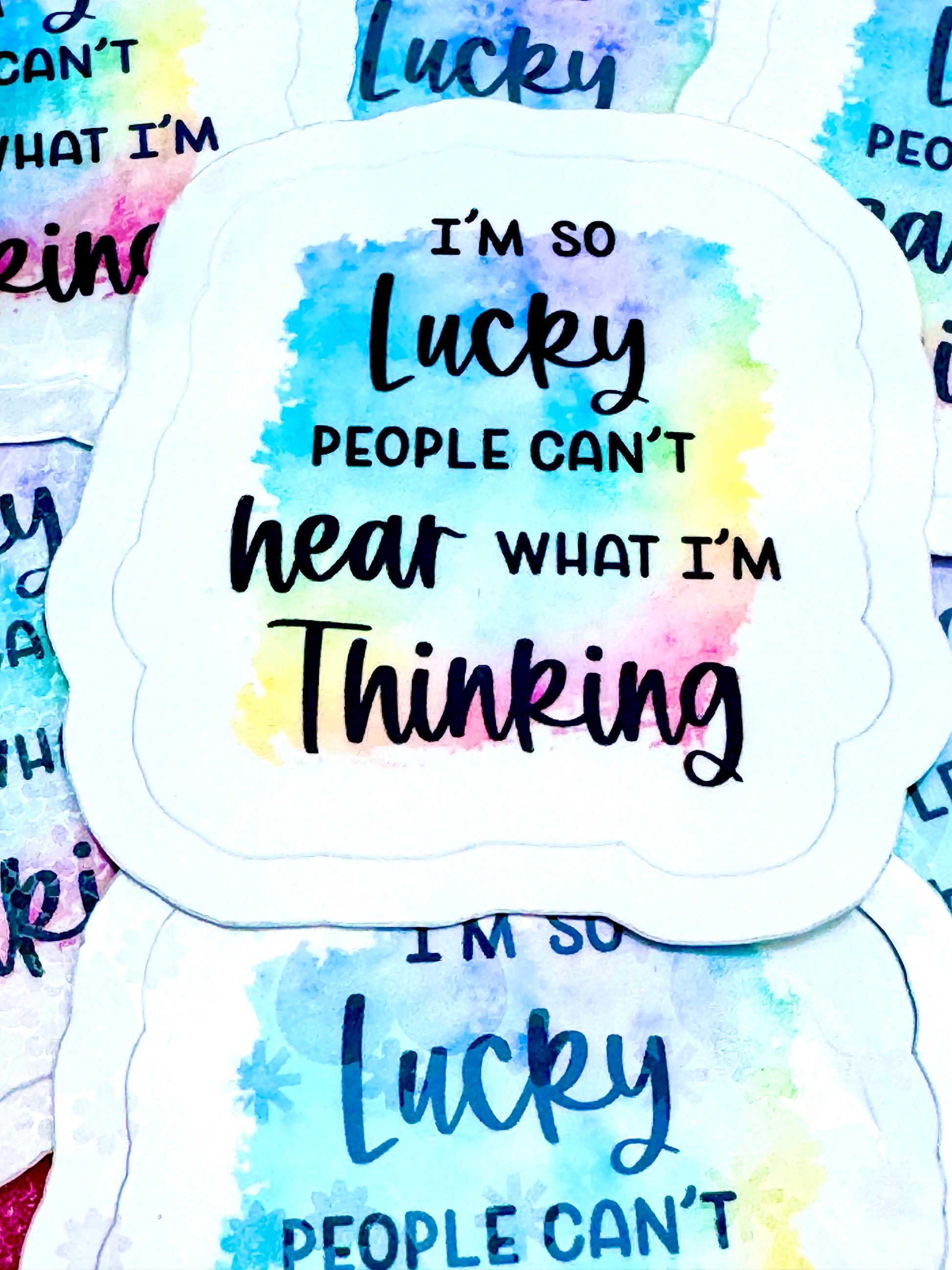 Vinyl sticker with a rainbow background and the phrase ‘Lucky People Don’t Hear What I’m Thinking’ in black font, photographed on a flat surface.