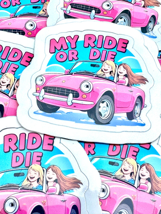 "Vinyl sticker featuring two girls, one blonde and one brunette, in a pink open-top car with the phrase ‘My Ride or Die’, photographed on a flat surface."