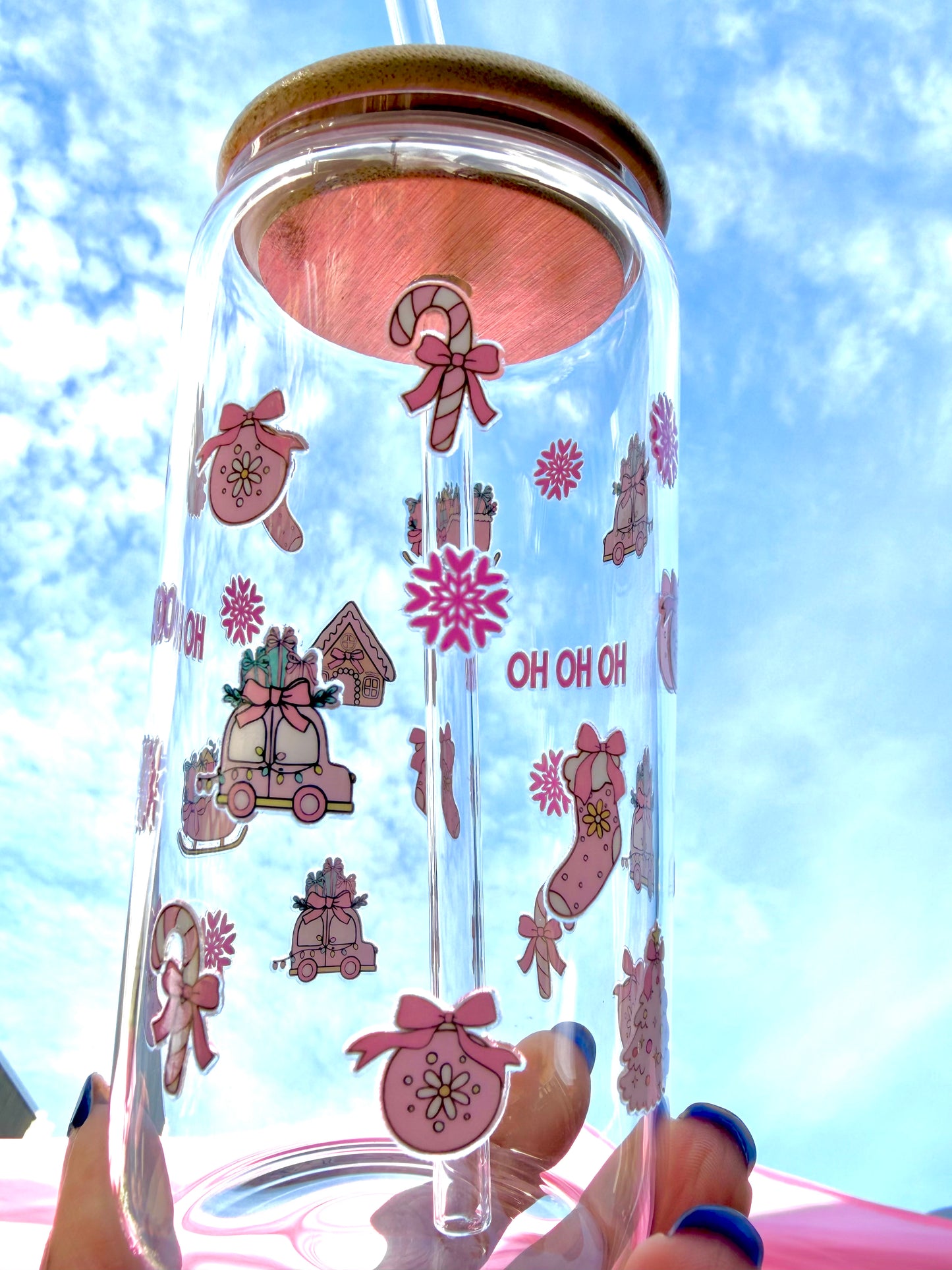 A reusable pink “HO HO HO” Christmas glass with bamboo lid and clear glass straw, decorated with candy canes, stockings, and gifts, held against a bright festive background.