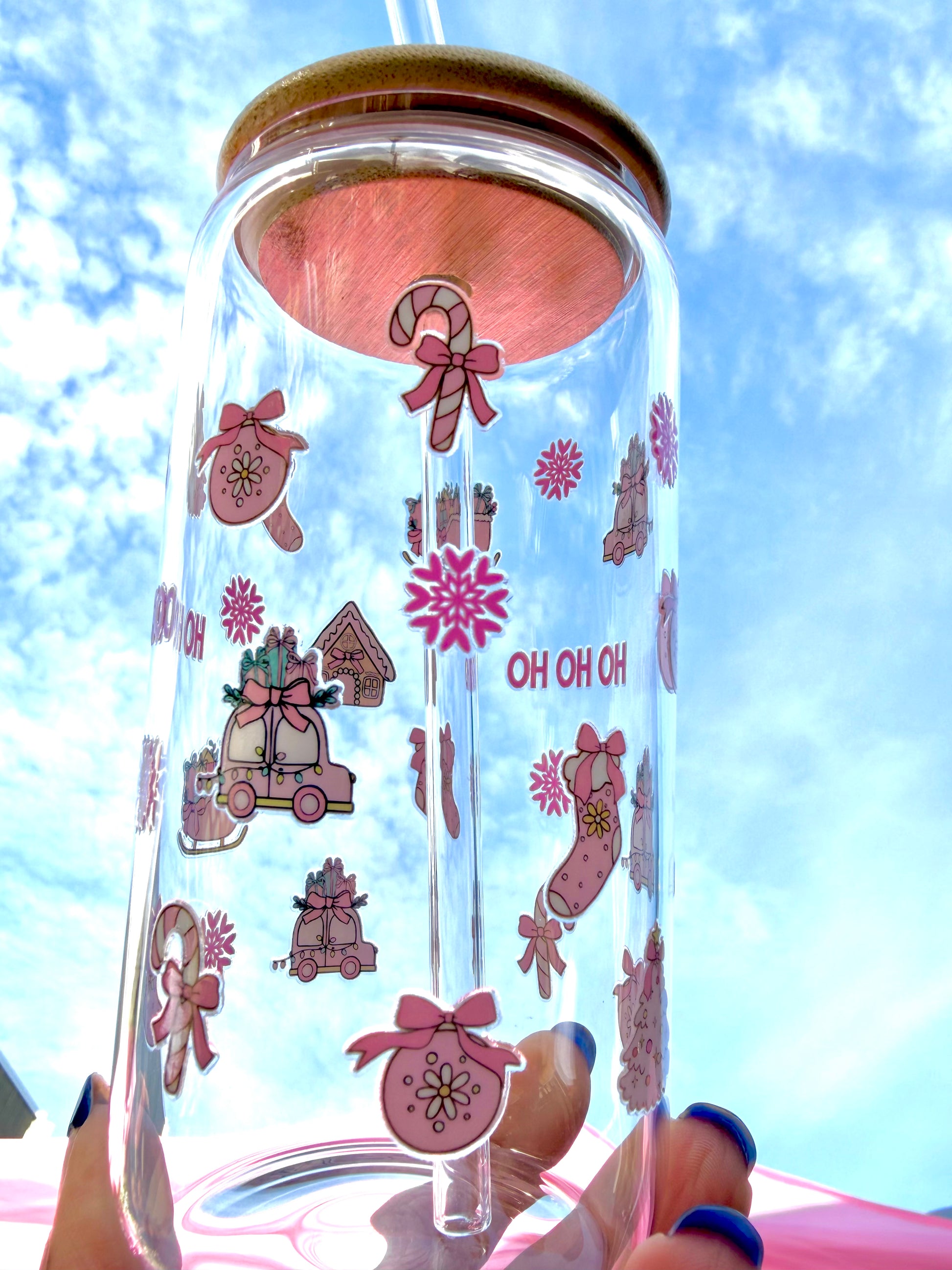 A reusable pink “HO HO HO” Christmas glass with bamboo lid and clear glass straw, decorated with candy canes, stockings, and gifts, held against a bright festive background.