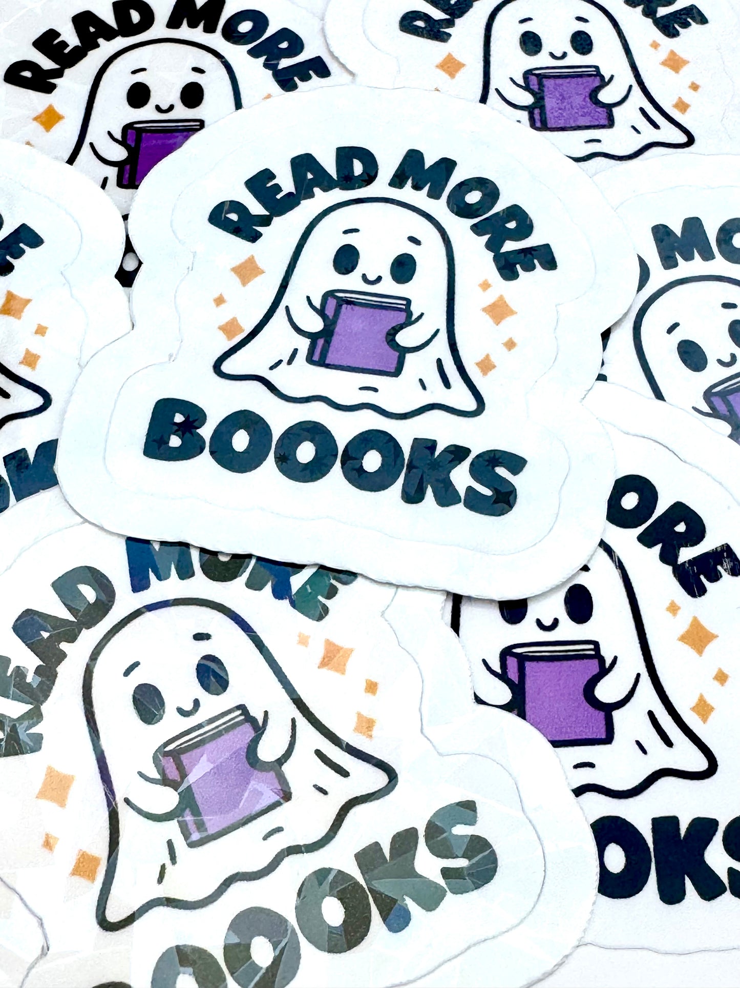 Vinyl sticker of a ghost holding a purple book with the phrase ‘Read More Booooks’ in black font, photographed on a flat surface.