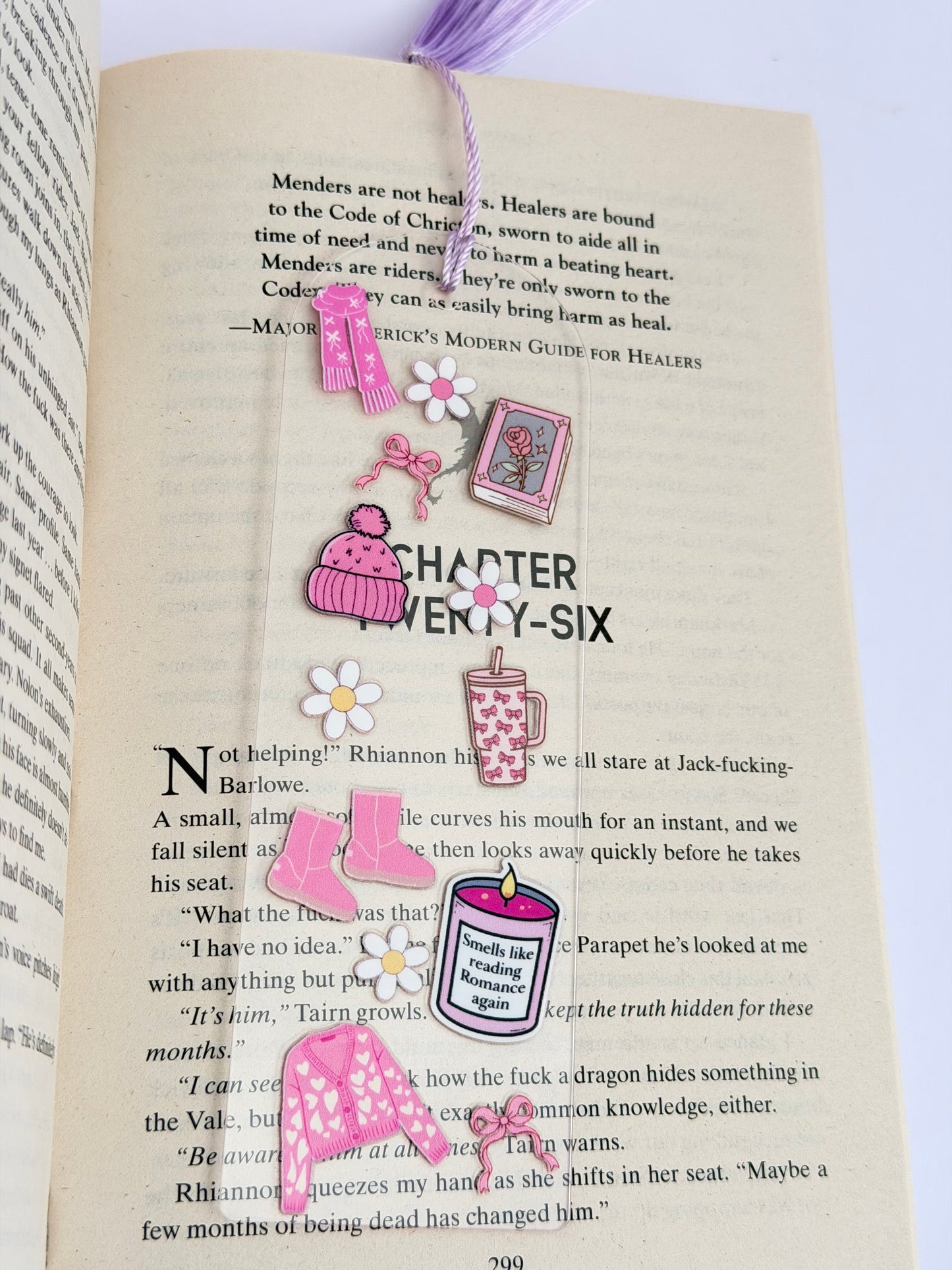 Close-up of The Romance Bookclub Acrylic Bookmark featuring cozy pink icons like a cardigan, boots, mug, and daisies, lying flat across an open page.