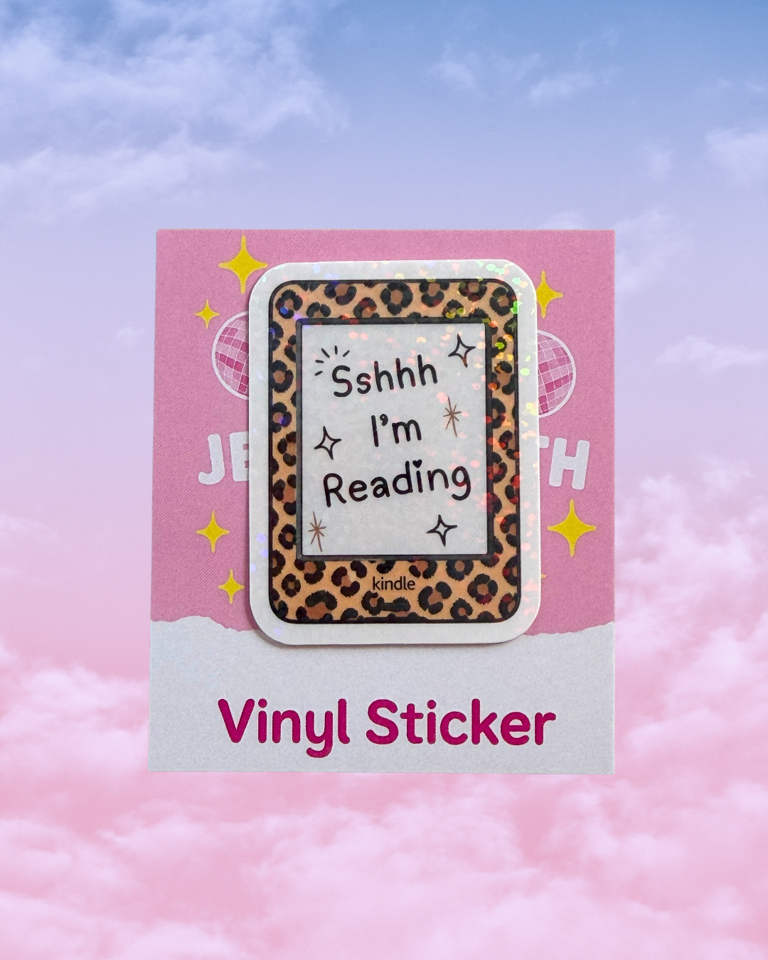 A rectangular vinyl sticker with a leopard print border e reader image and a white center reading “Sshhh I’m Reading,” displayed on a pink backing card against a pastel pink-and-blue cloudy sky background.