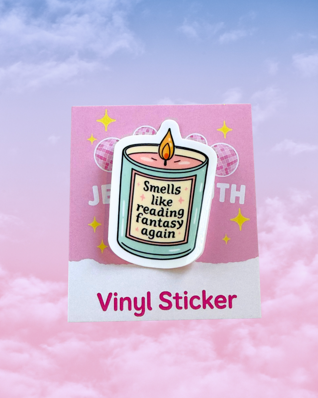 A die-cut vinyl sticker of a candle with the text 'Smells like reading fantasy again' displayed on a pink backing card that reads 'Vinyl Sticker', set against a pastel pink and blue cloudy background.