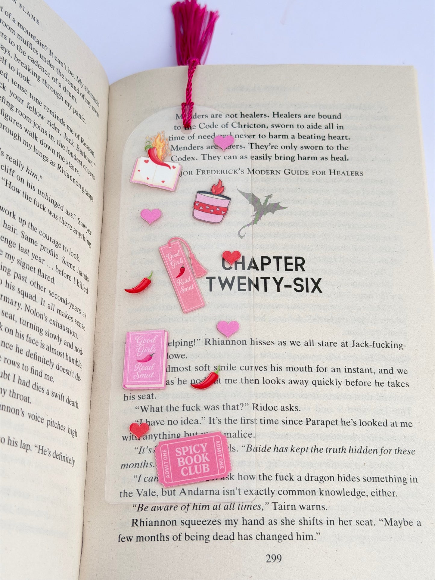 A clear acrylic bookmark with pink and red spicy book-themed designs, including hearts, books, and chilli peppers, lying across an open book with a pink tassel at the top.