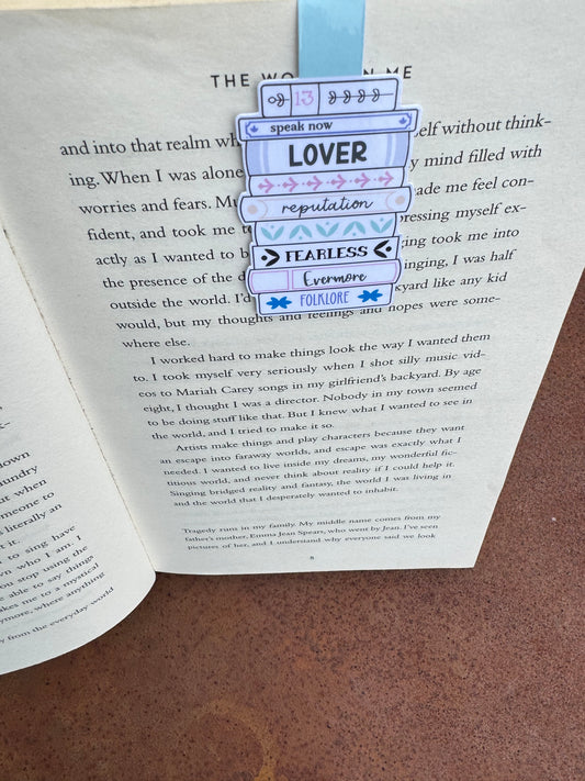Magnetic bookmark is sat on the page of a book the design is a stack o books with swift album titles on the books