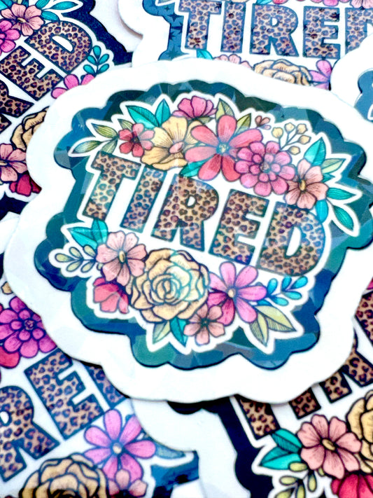 vinyl sticker with the word ‘TIRED’ in leopard print font, surrounded by floral illustrations, photographed on a flat surface