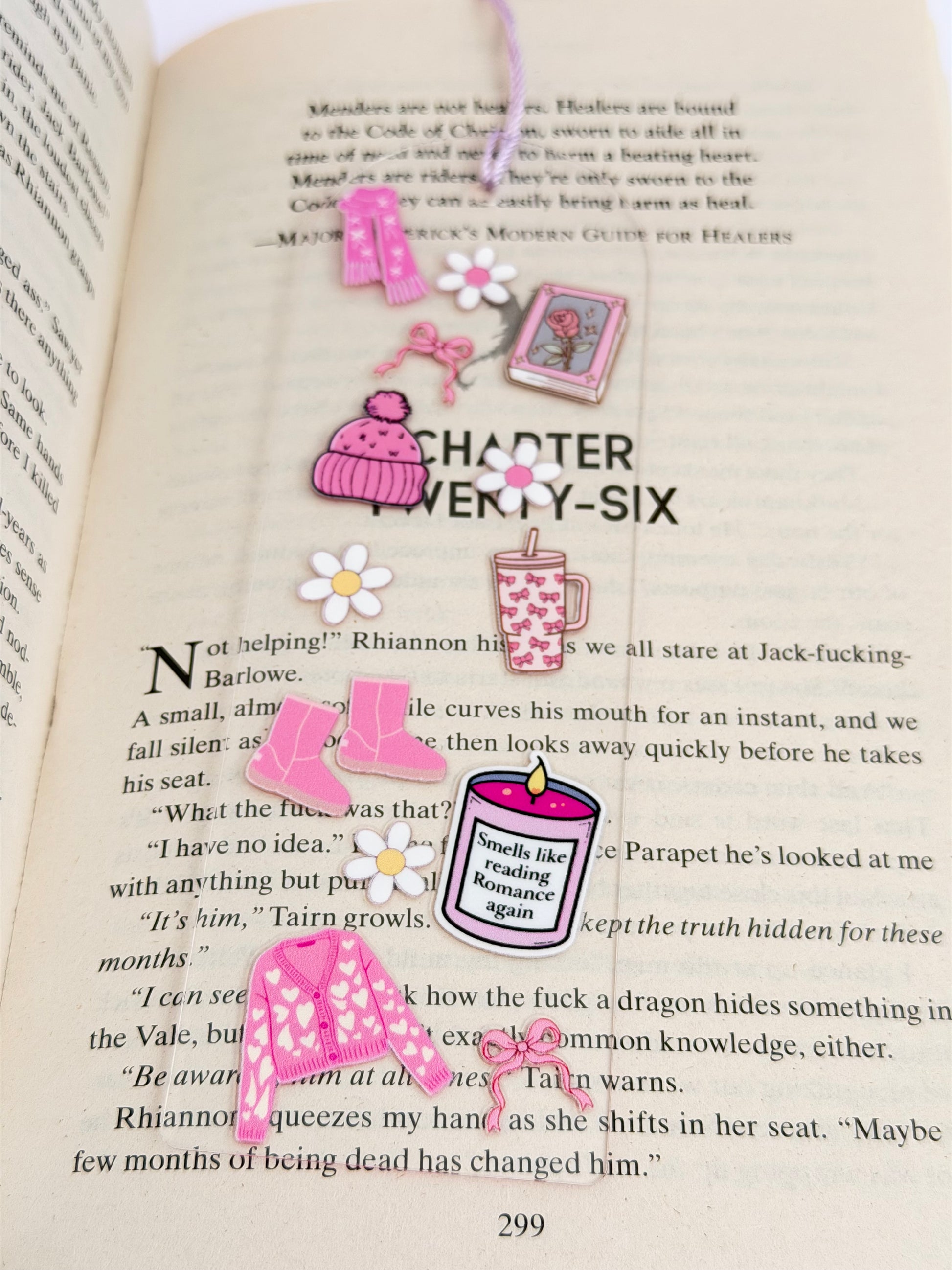 A clear acrylic bookmark with pink and white romance-themed designs including daisies, bows, and a candle reading “Smells like reading romance again,” shown on an open book with a lilac tassel.