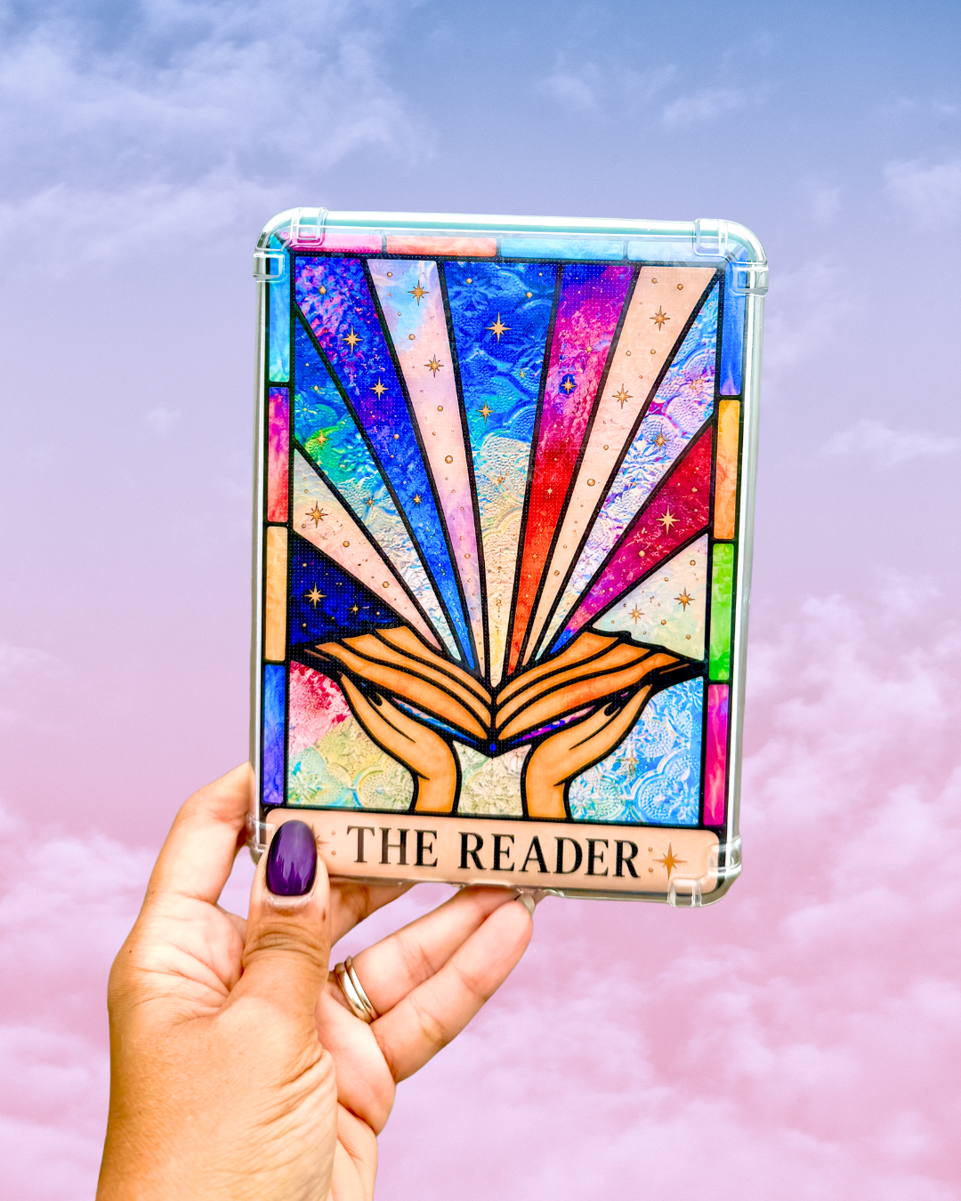 A hand with purple nail polish holds a clear-edged tarot card kindle insert labeled 'THE READER' against a pastel pink and blue cloudy sky background. The box features a stained-glass-style illustration of open hands holding a book with multicolored rays and stars.